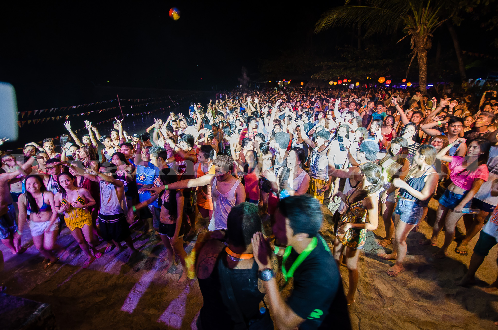 A massive crowd of people dancing and celebrating at a nighttime beach party, illuminated by colorful lights and floating lanterns against a dark sky, with a palm tree visible at the right edge.