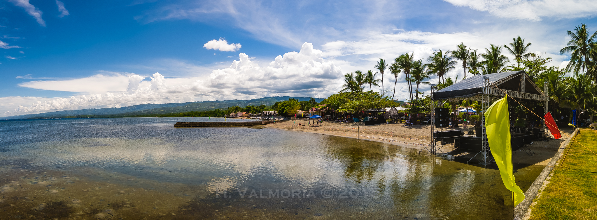A wide panoramic view of a Philippine beachfront event venue with a metal concert stage and colorful flags on the right, palm trees lining the shore, calm reflective water on the left, and dramatic white clouds over distant green mountains.