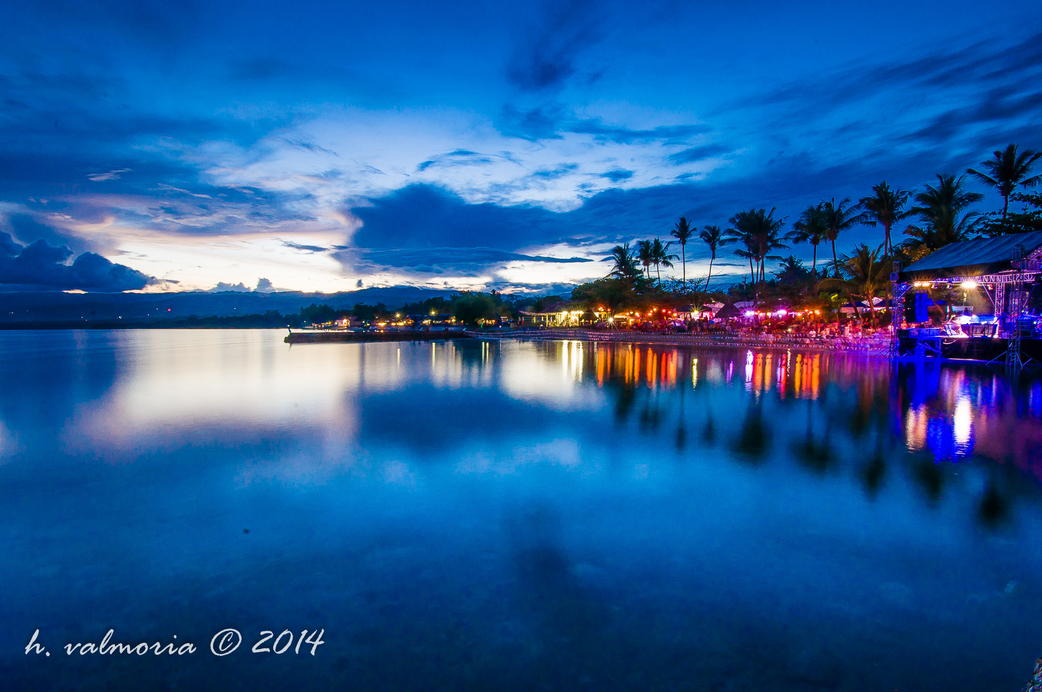 A twilight panorama of a Philippine bay at blue hour, with colorful festival lights and palm tree silhouettes reflecting on glassy water while dramatic clouds stretch above a fading sunset on the horizon.