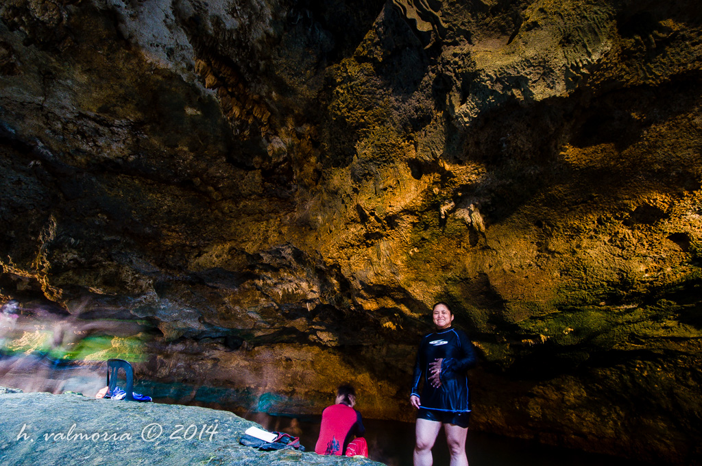 A woman in a black swimsuit stands smiling inside a dramatic limestone cave, with warm golden light illuminating the textured rock ceiling above. A second person sits in the background near a dark underground pool.