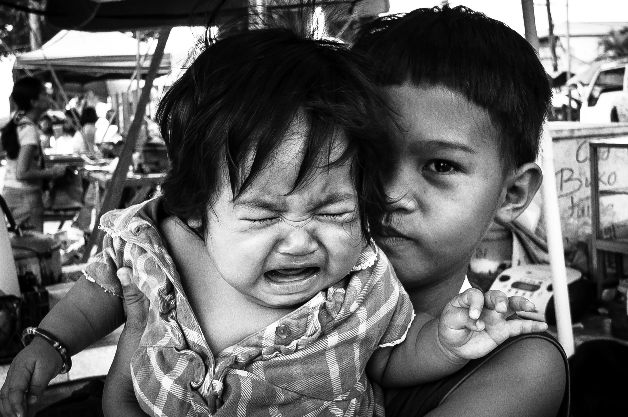 A black and white close-up of two young children at a busy market, with an older boy holding a crying baby against his cheek, looking directly at the camera with a calm, knowing expression.