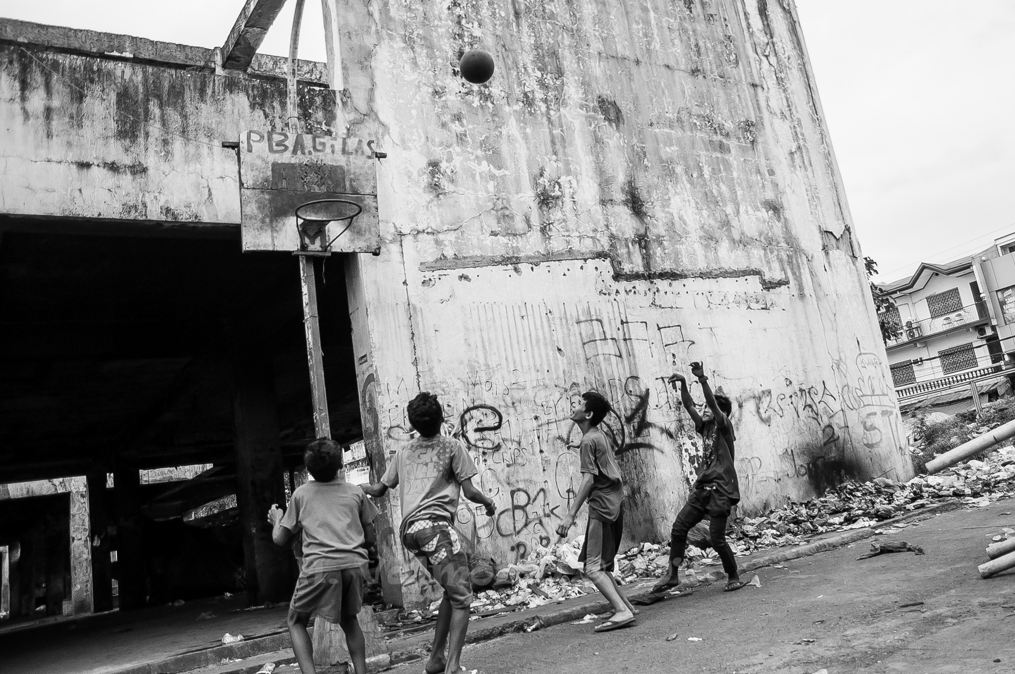 A group of children playing basketball against a weathered, graffiti-covered concrete wall in an urban Southeast Asian setting in black and white.