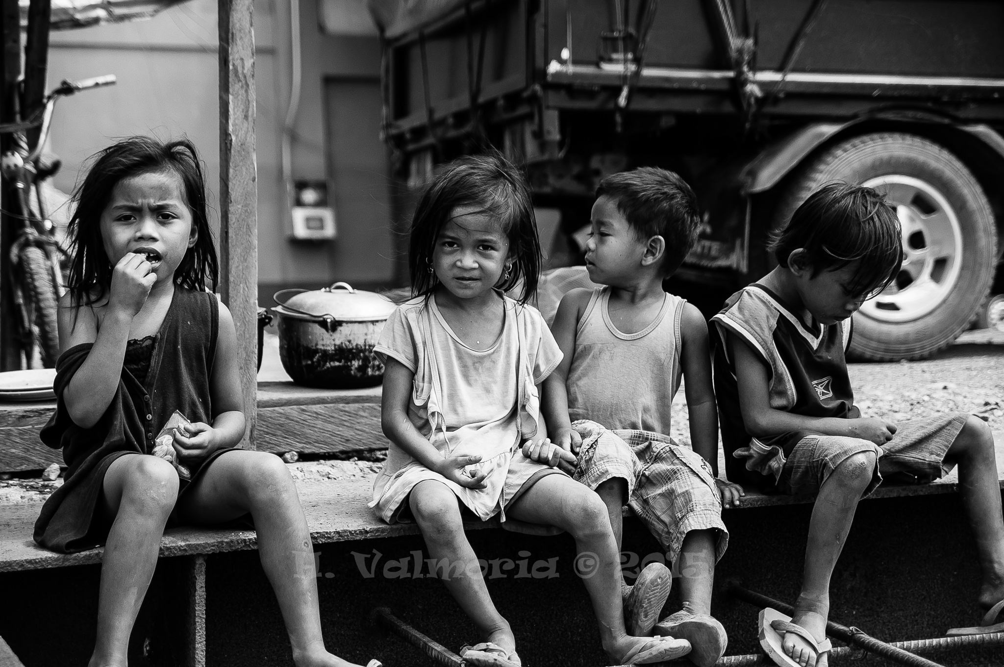 Four young children sit together on a low ledge in a street setting in black and white, one girl eating while another gazes directly into the camera.