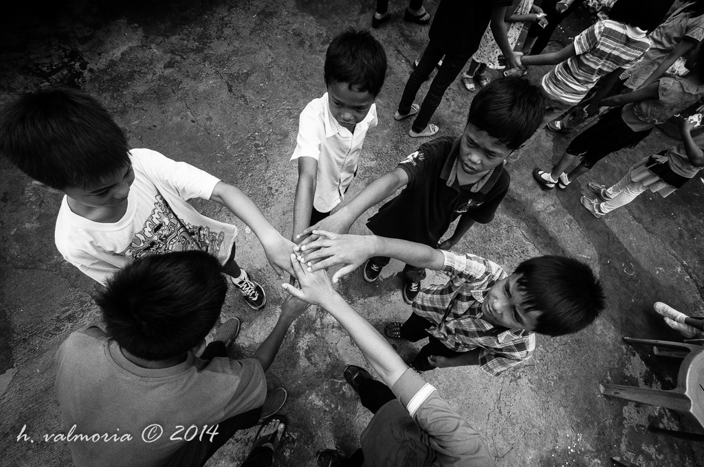 A high-angle black and white photograph of a group of children stacking their hands together in a circle, viewed from above, with onlookers visible at the edges of the frame.