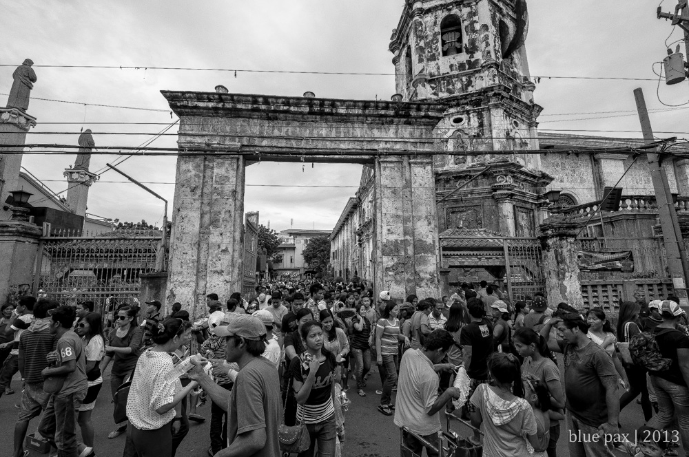 A dense crowd of Filipino people streaming through the stone gateway of a historic Spanish colonial church in black and white, with a weathered bell tower rising behind them under an overcast sky.