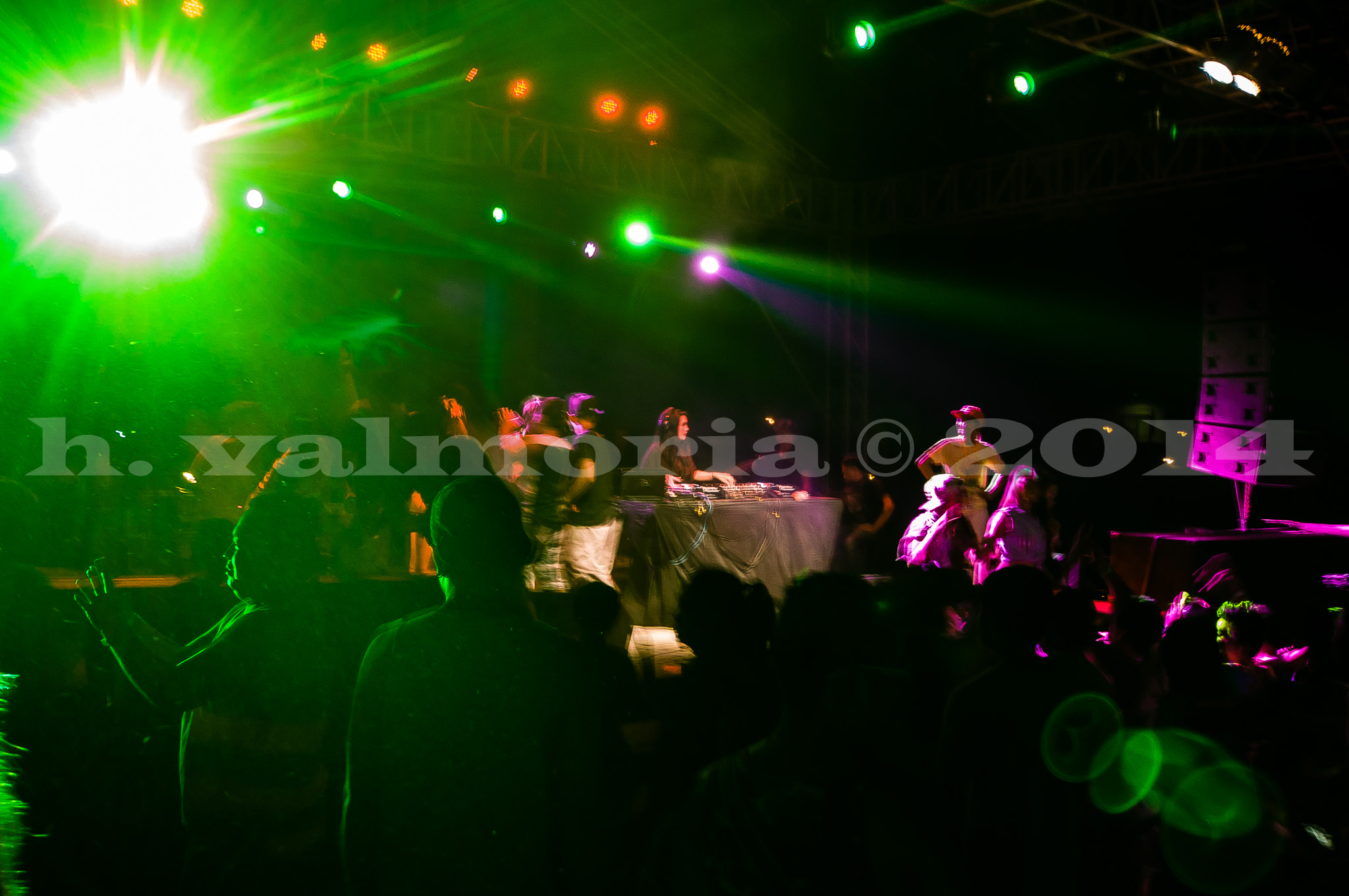 A crowd of silhouetted figures watch a performer on a stage bathed in vivid green and pink stage lights. The long-exposure shot captures motion blur and the electric atmosphere of an outdoor nighttime event.