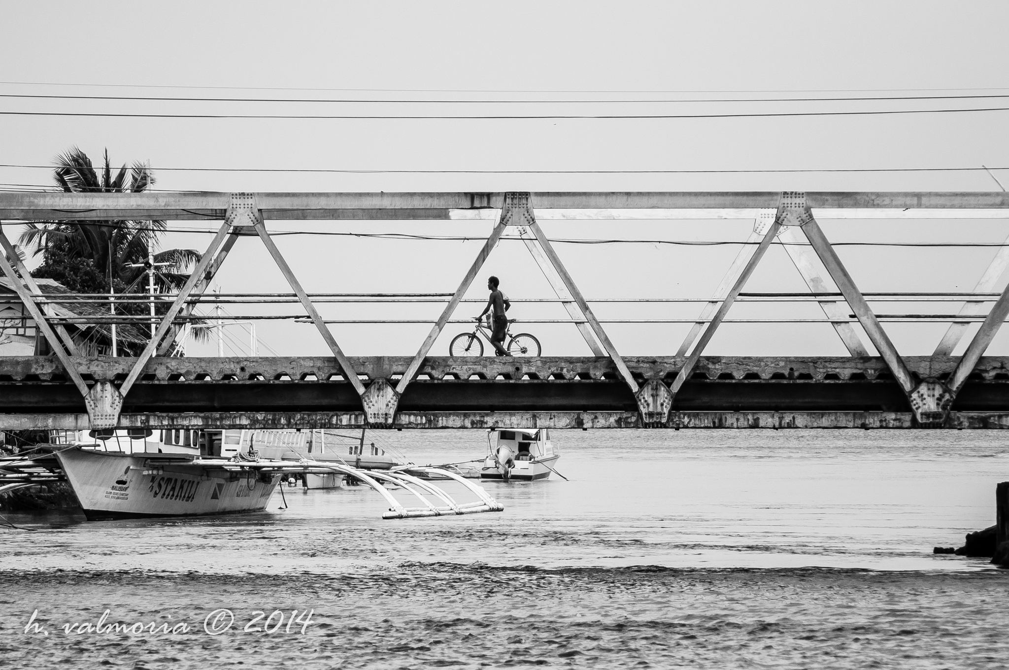 A lone cyclist silhouetted against a steel truss bridge in black and white, with traditional Filipino outrigger bangka boats moored in the water below.