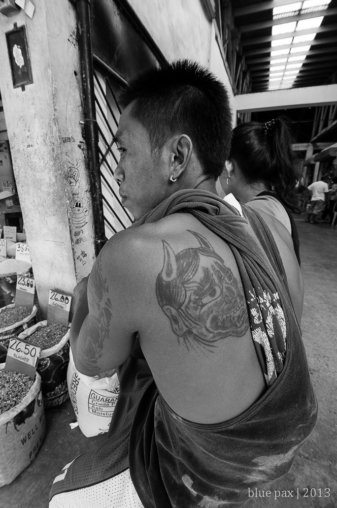 A young man seen from behind at a market stall in black and white, his sleeveless shirt revealing a large demon oni tattoo across his shoulder, with sacks of grain and price tags visible.