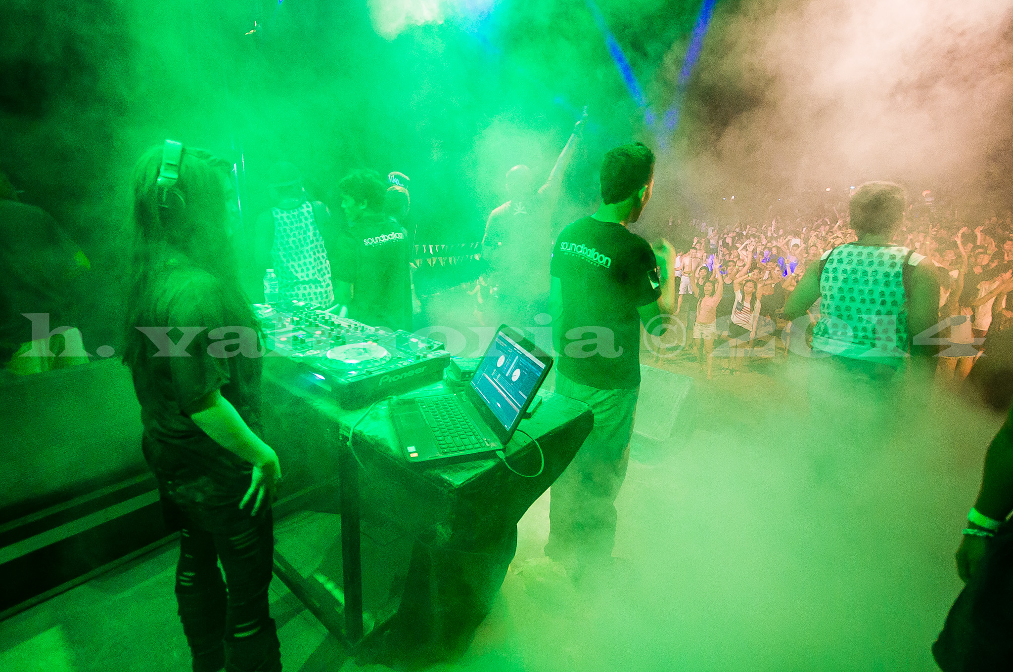 DJ crew works at a Pioneer mixer and laptop on a stage bathed in intense green stage smoke, while a large crowd dances in the background, giving a performer's-eye view of the packed dancefloor.