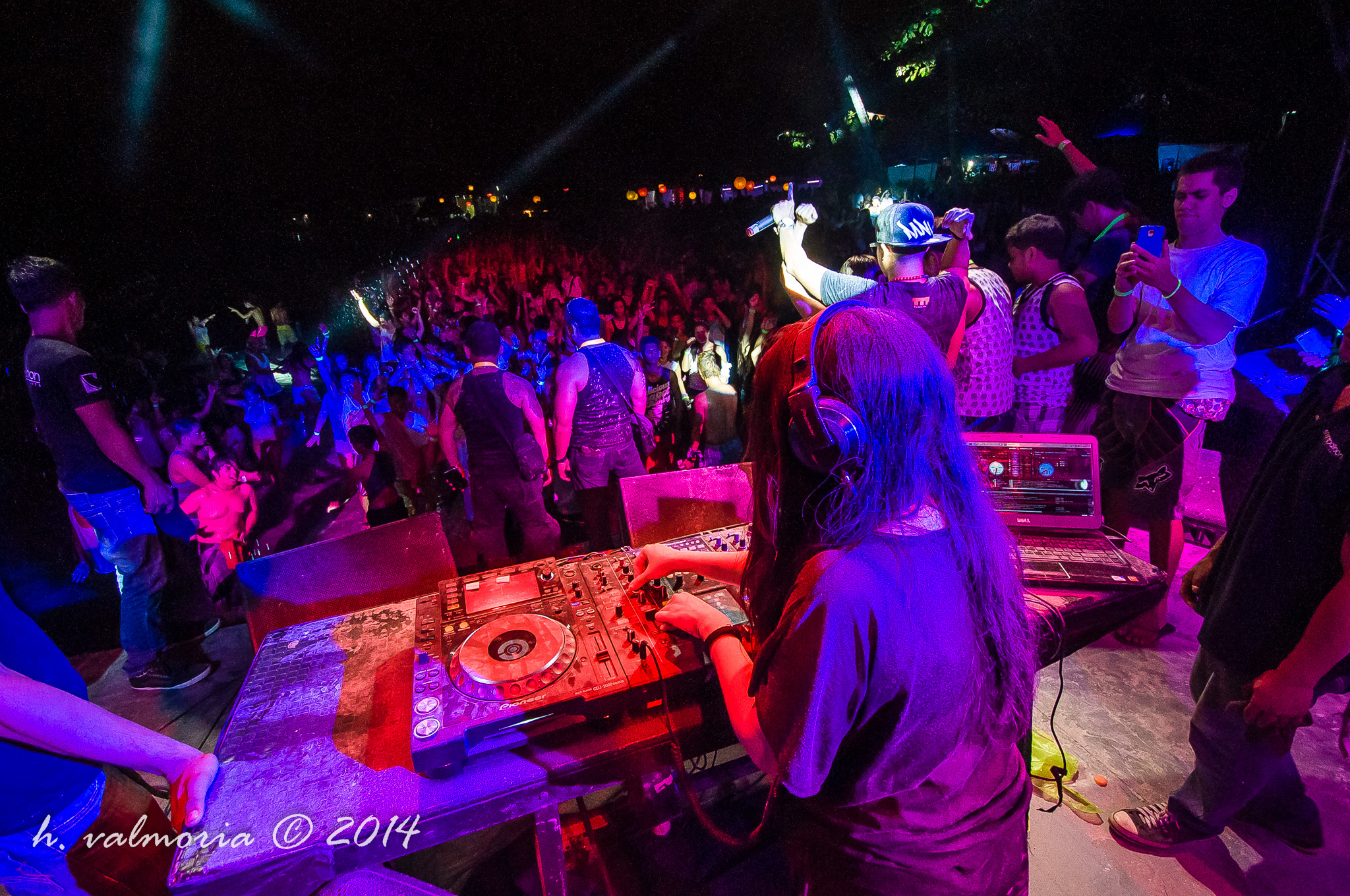 A DJ performs at a packed outdoor concert, viewed from behind the Pioneer mixer and CDJs as dramatic purple and red stage lights wash over a dense crowd stretching into the darkness.