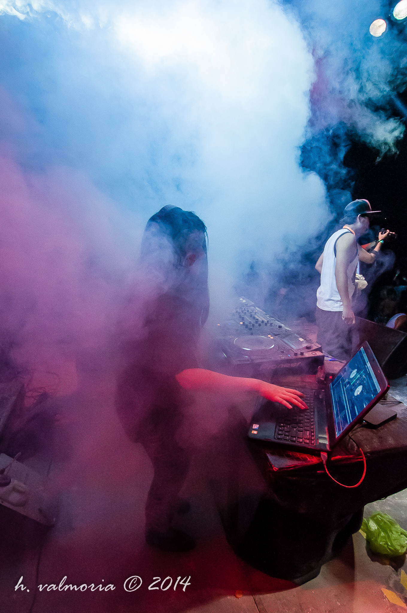 A DJ hunches over turntables and a laptop at an outdoor event, nearly swallowed by thick blue and pink smoke machine haze, while a performer with a microphone stands nearby under stage lights.