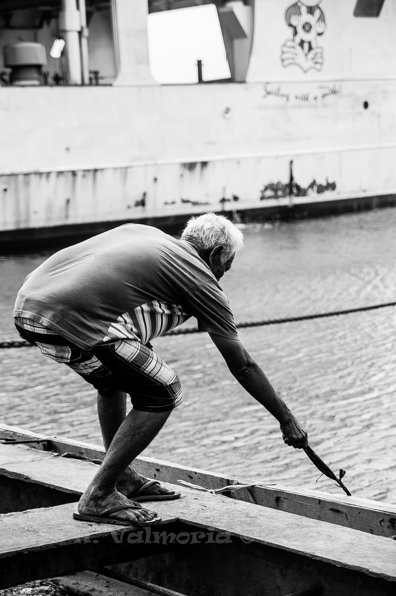 An elderly man leans forward on a wooden dock in black and white, gripping a tool with weathered hands, with a large vessel and rippling water visible in the background.
