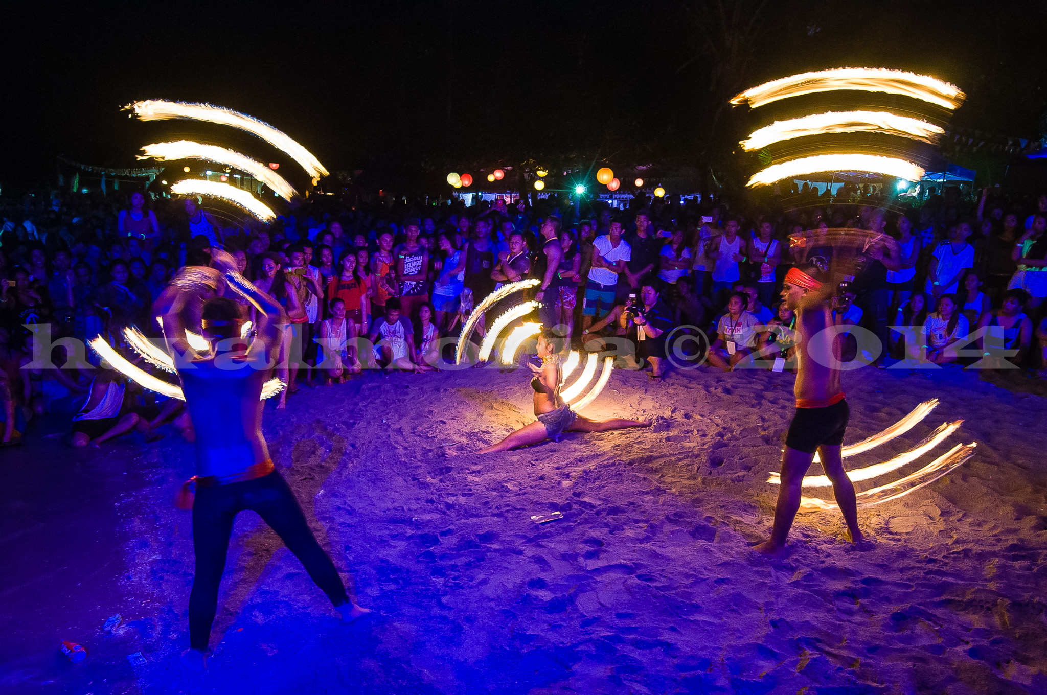 Fire dancers spin glowing poi on a sandy beach at night, creating sweeping arcs of light, while a large crowd watches under blue and purple ambient lighting.