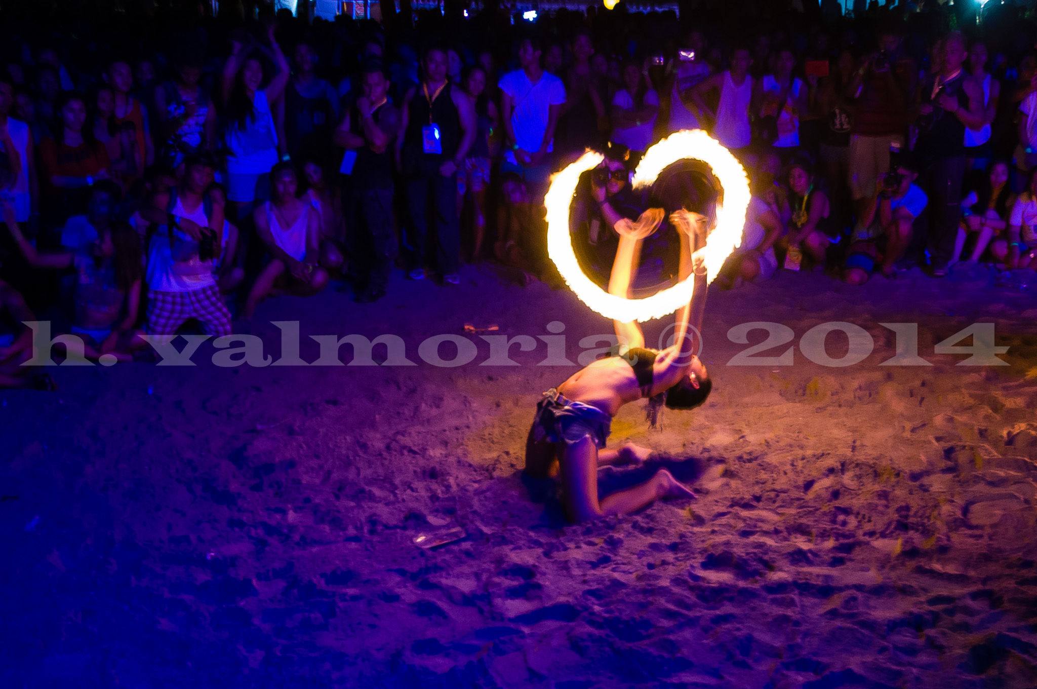 A fire dancer kneels on a sandy beach at night, arching backward while spinning a large flaming ring above their body. A dense crowd of spectators watches from the background, bathed in blue and purple light.