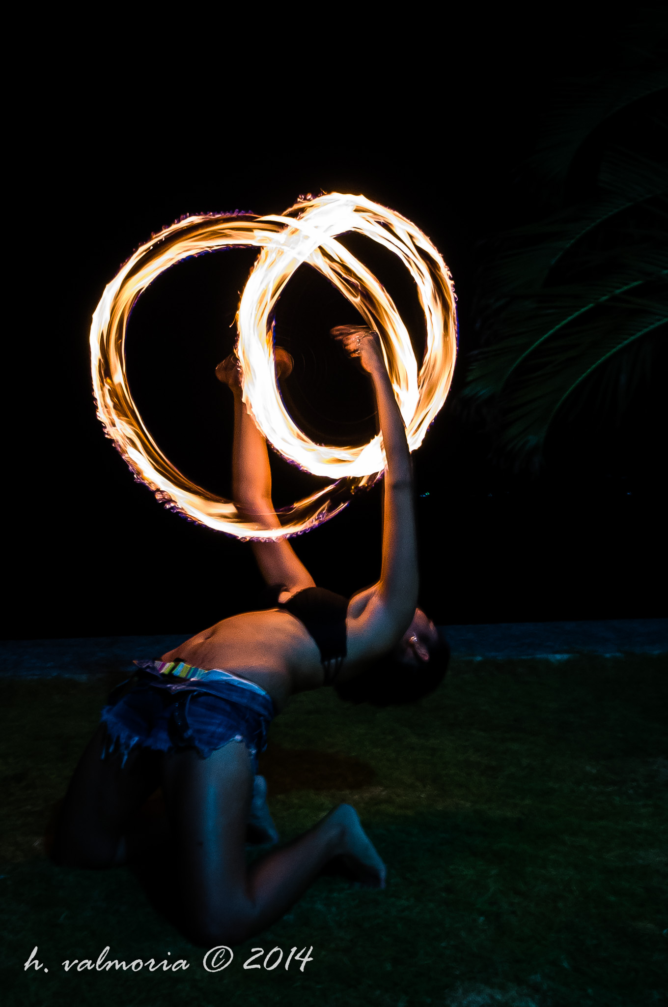 A fire dancer kneels on grass at night, spinning twin fire poi that trace glowing circular arcs of light against a pitch-black background, silhouetted by the warm orange flames.