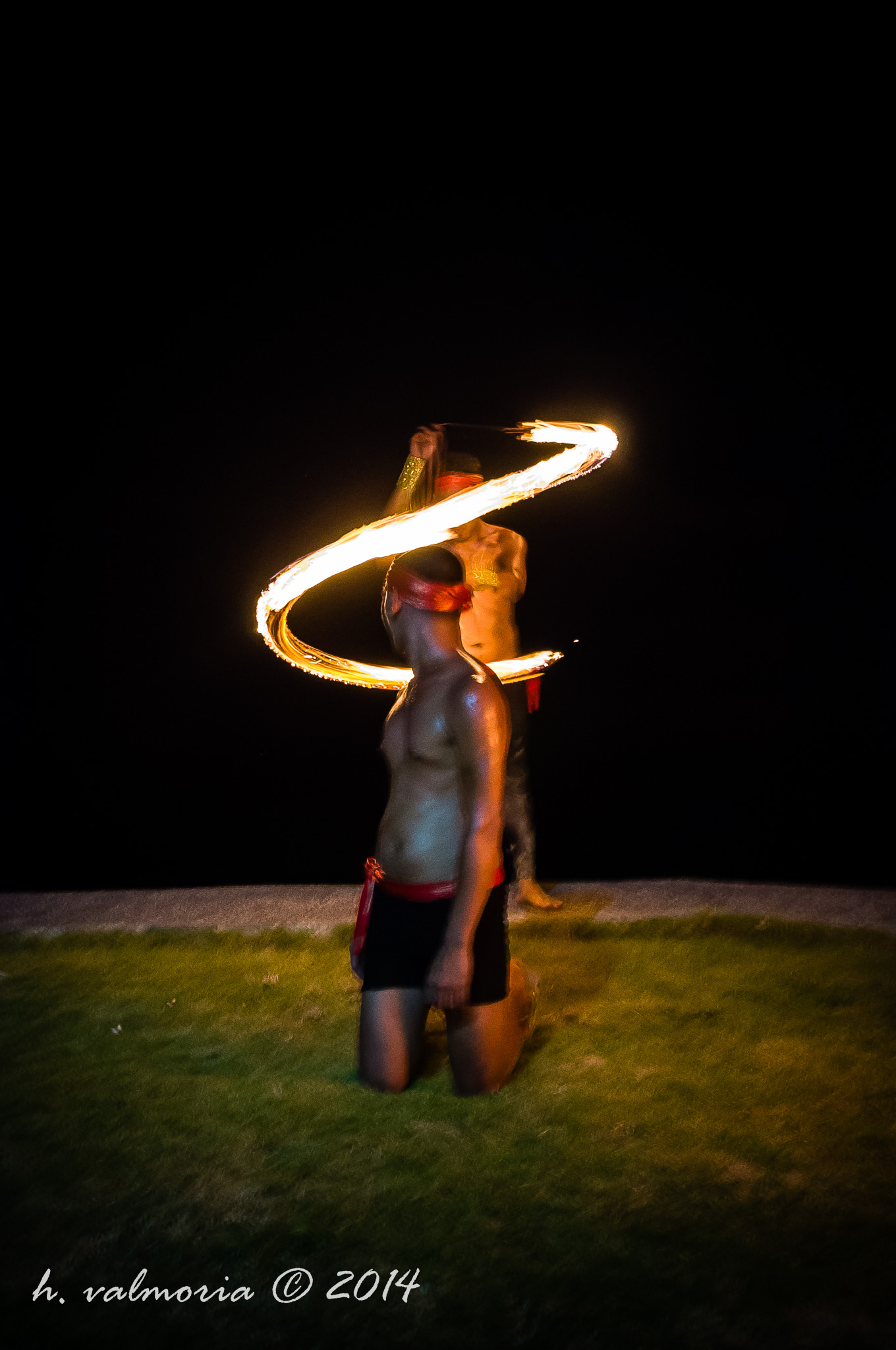 A shirtless fire dancer kneels on grass at night while a second performer spins a fire tool behind them, leaving a glowing spiral trail of flame against a pitch-black sky.