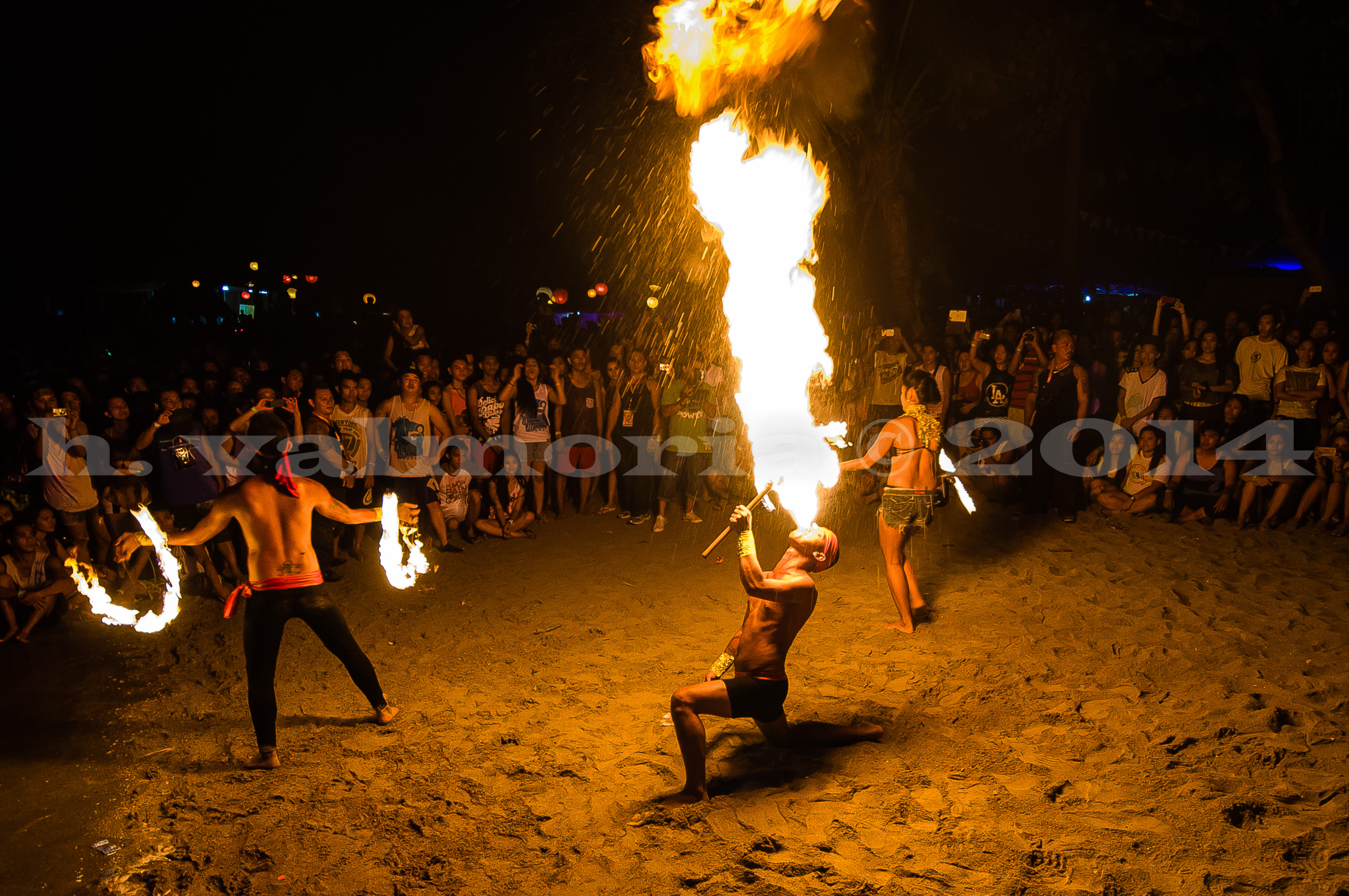 Three fire performers entertain a large crowd on a dark beach at night, with one performer breathing a tall column of flame while two others spin fire poi, all illuminated by the warm orange glow of the flames.