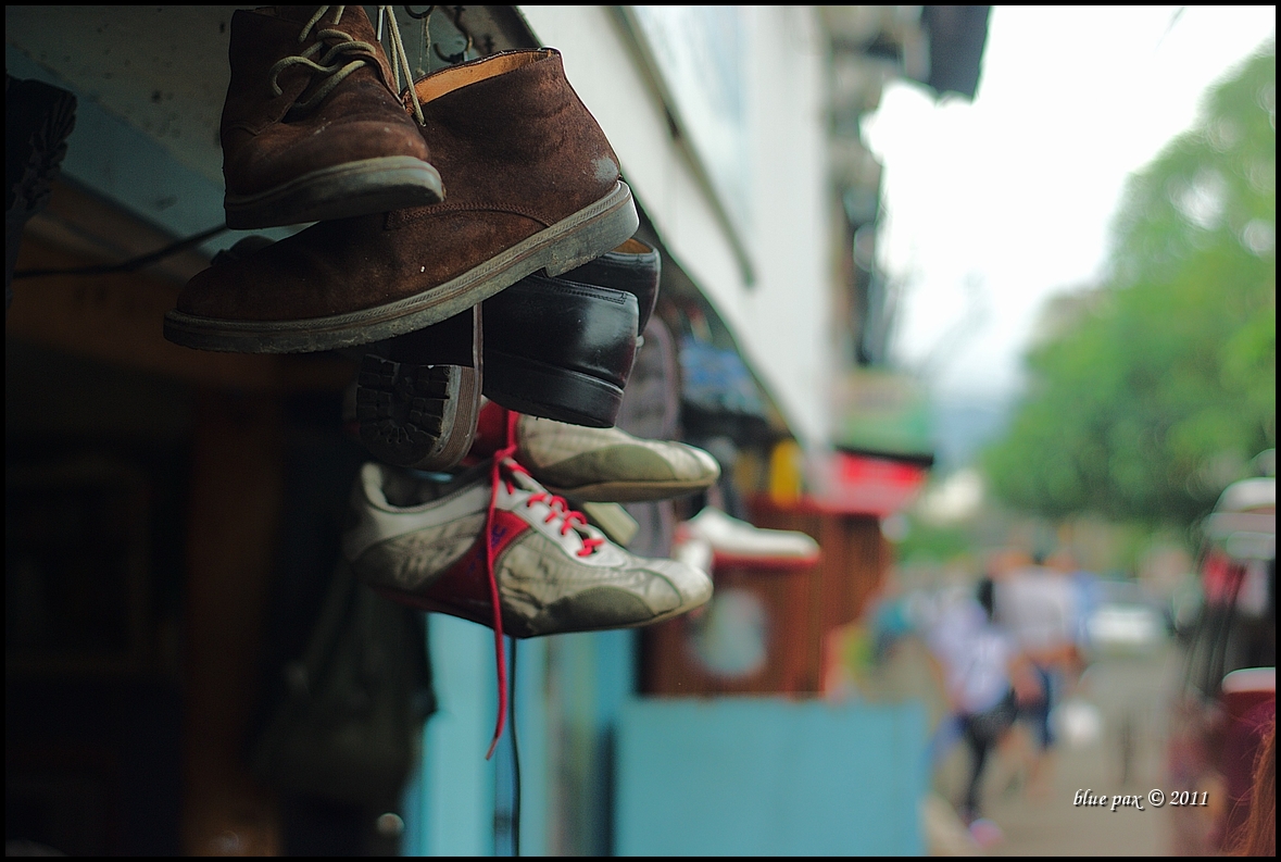 Several pairs of worn shoes hang from a storefront overhang in the foreground, with a blurred busy street market scene stretching into the background.