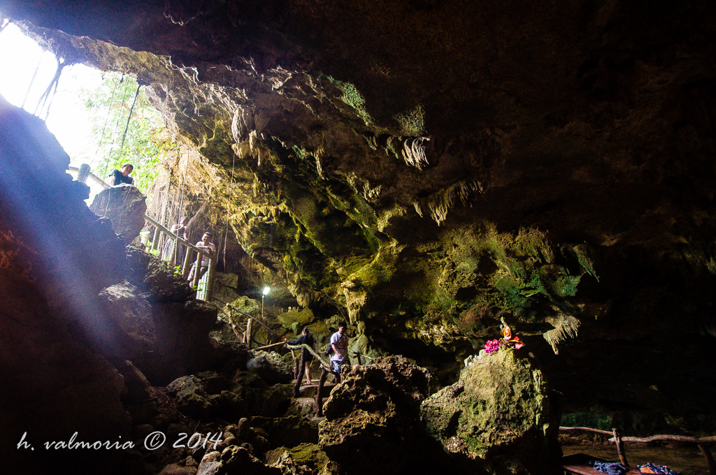 Dramatic shafts of light pierce through a cave opening in the Philippines, illuminating figures descending wooden walkways and a small religious shrine placed on a mossy rock formation in the cavern below.