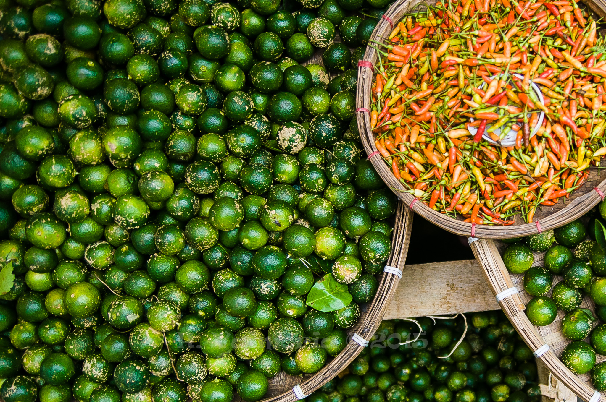 An overhead view of a market stall showing woven baskets overflowing with small green limes and a round basket filled with vibrant red and orange chili peppers.
