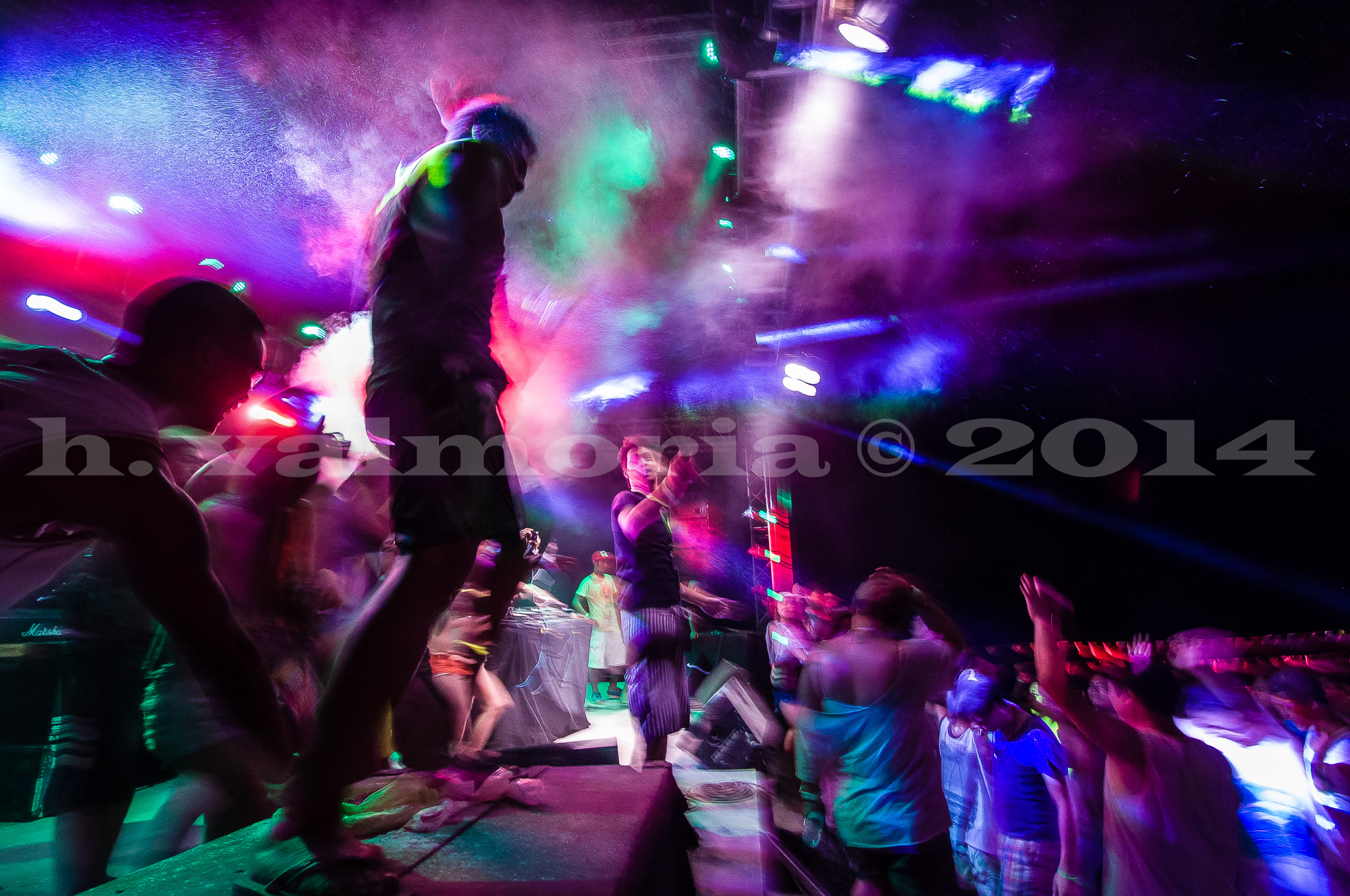 A crowded indoor concert scene illuminated by vivid purple, green, and red stage lights, with dancers in motion blur and colored smoke drifting through the air above the crowd.