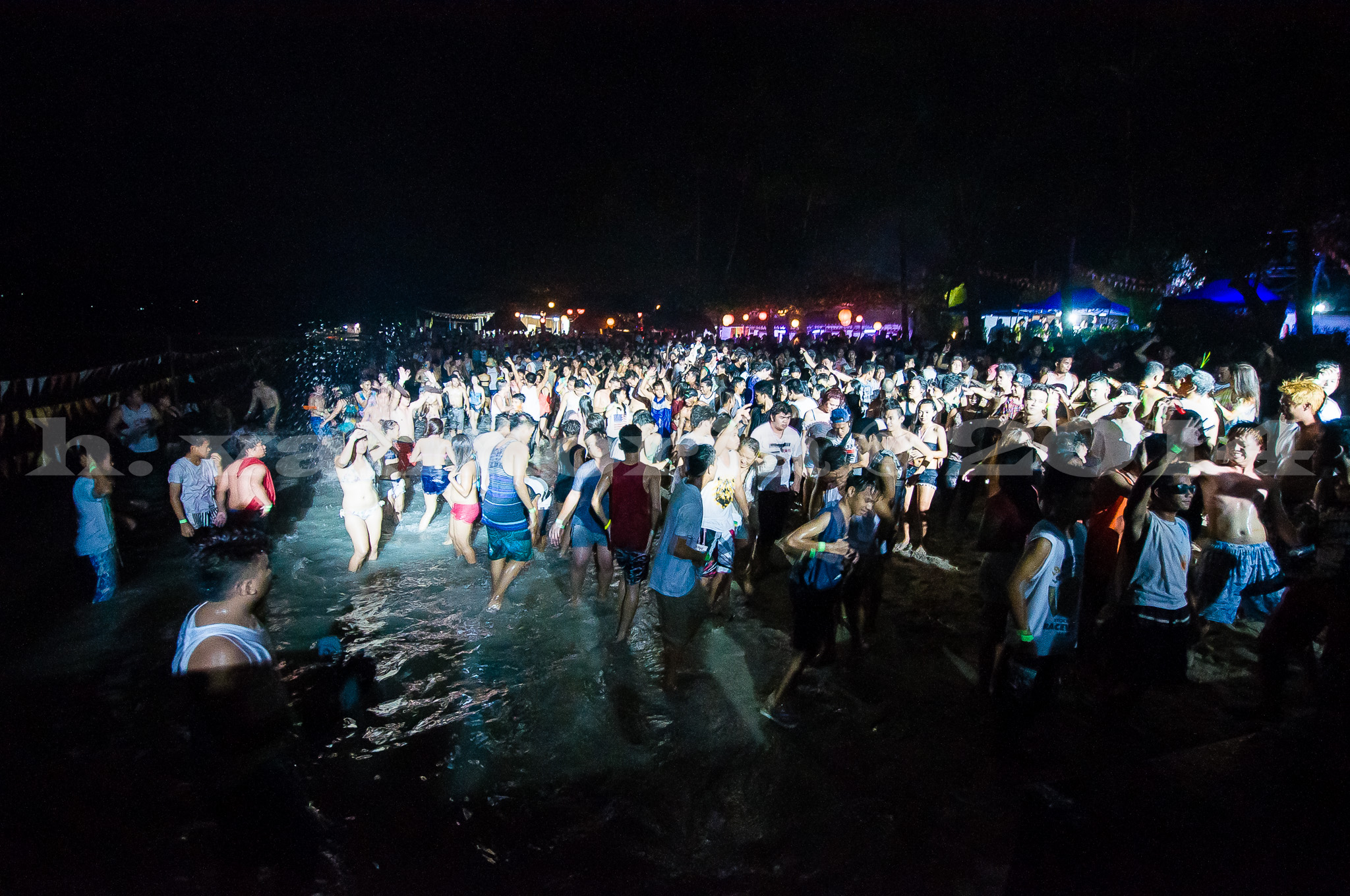 A massive crowd of people wade and gather in shallow beach water at night, lit by artificial lights against a dark sky, with festive lights and stalls visible along the shoreline in the background.
