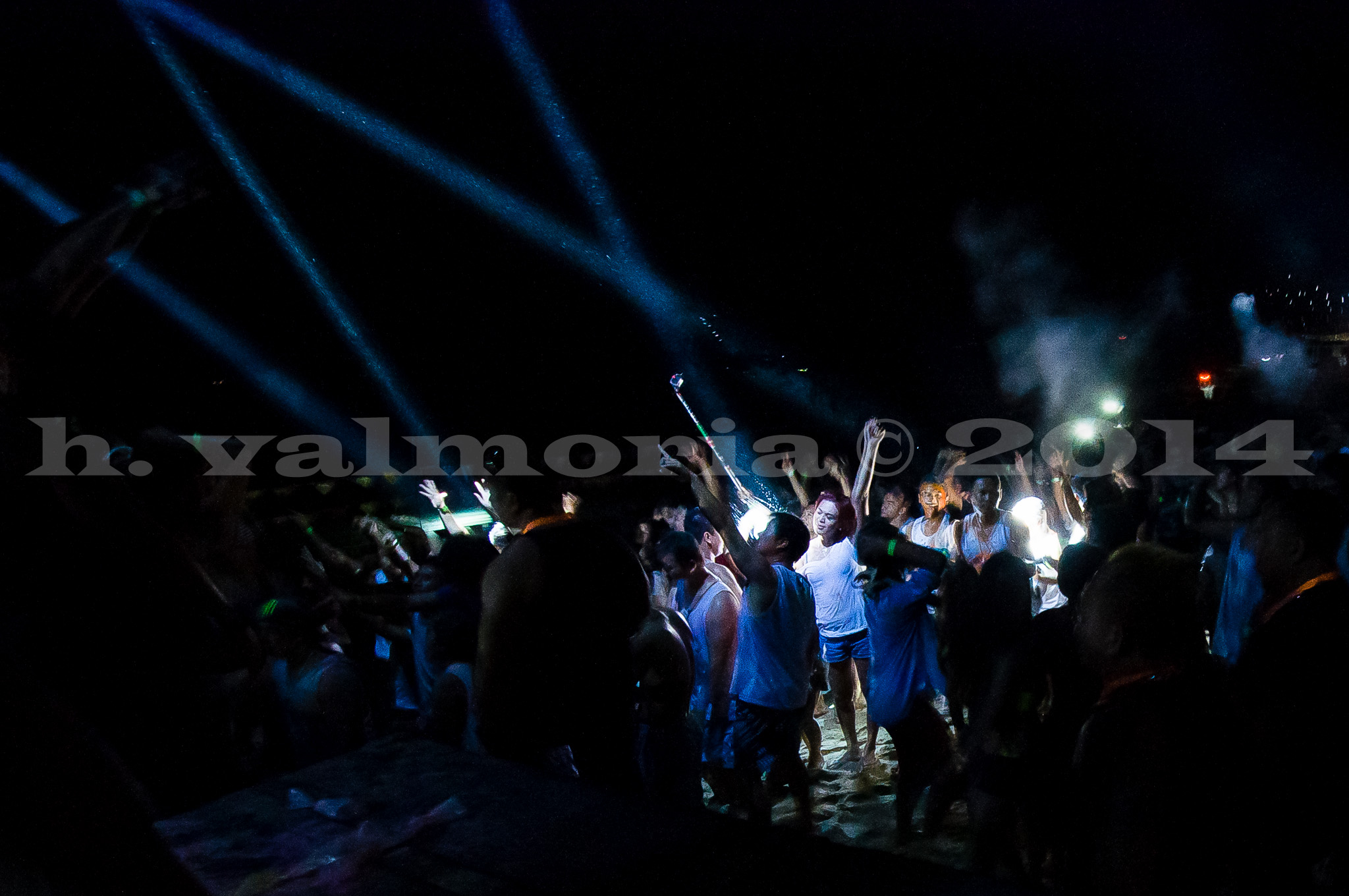A crowd of people dancing on a beach at night, illuminated by sweeping blue stage lights and handheld glowing devices. The dark sky is cut through with bright light beams as revelers raise their arms in celebration.