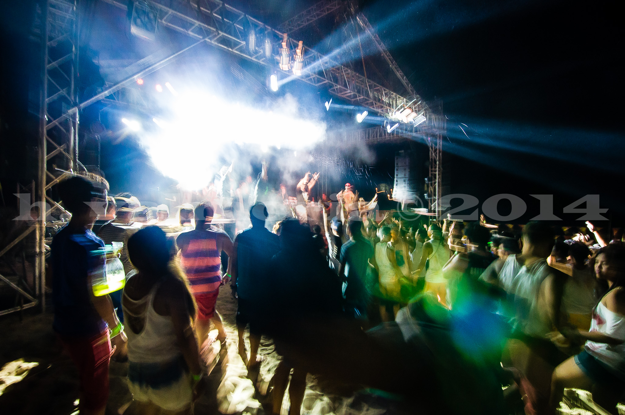 A large crowd gathered at an outdoor concert at night, facing a brightly lit stage with truss rigging and powerful spotlights cutting through smoke, with silhouetted figures dancing and celebrating under dramatic stage lighting.