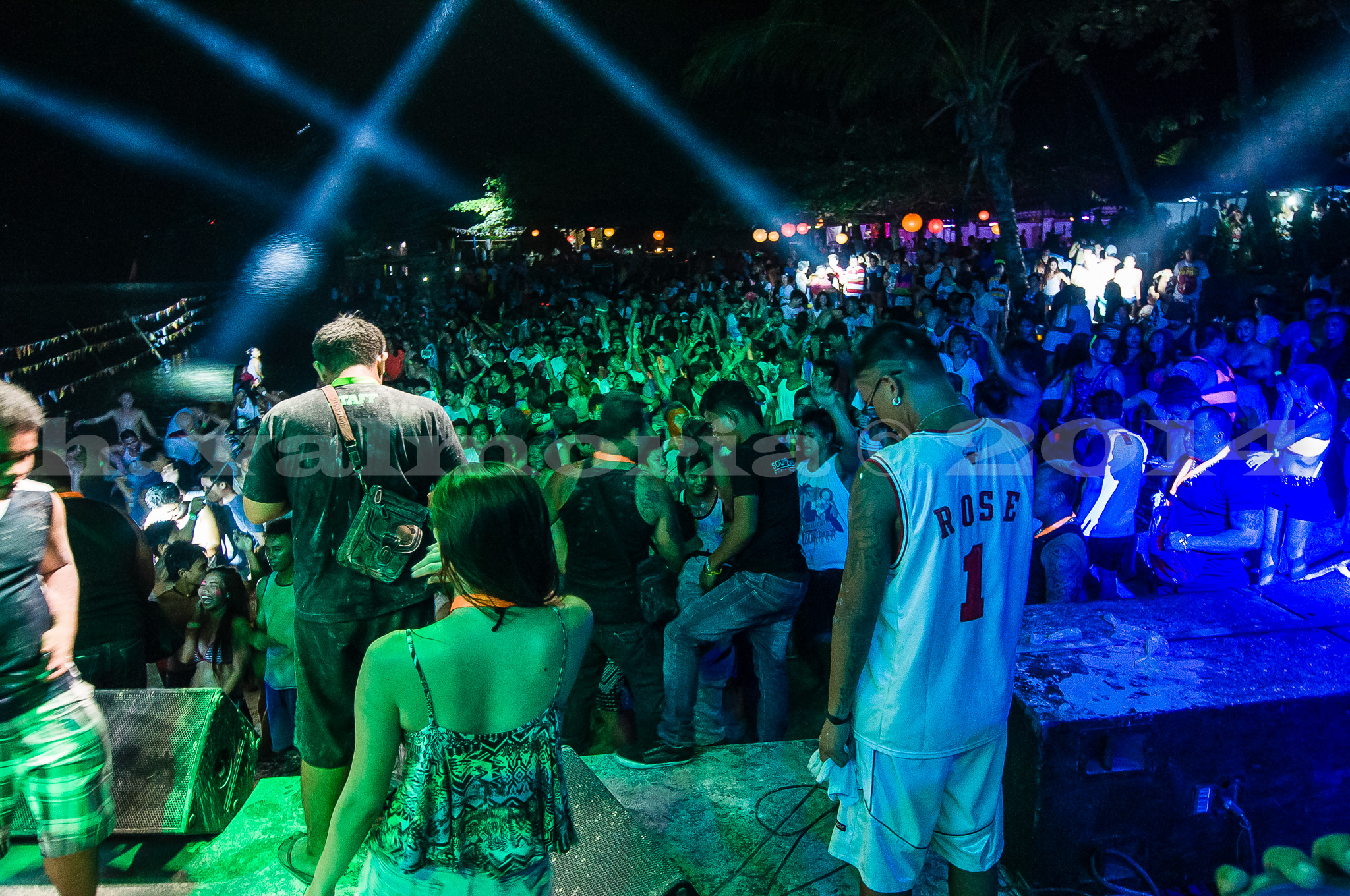 A large crowd of concertgoers fills an outdoor festival venue at night, illuminated by blue and green stage lights with dramatic light beams cutting through darkness while performers and attendees stand near the stage.