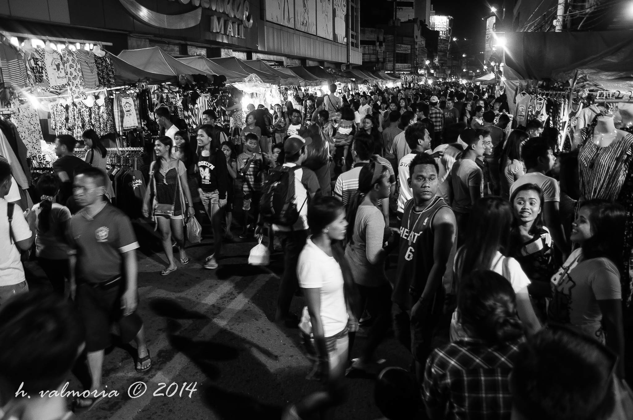 A dense crowd of shoppers fills a bustling outdoor night market in the Philippines in black and white, with clothing stalls lining both sides and neon lights glowing in the background.