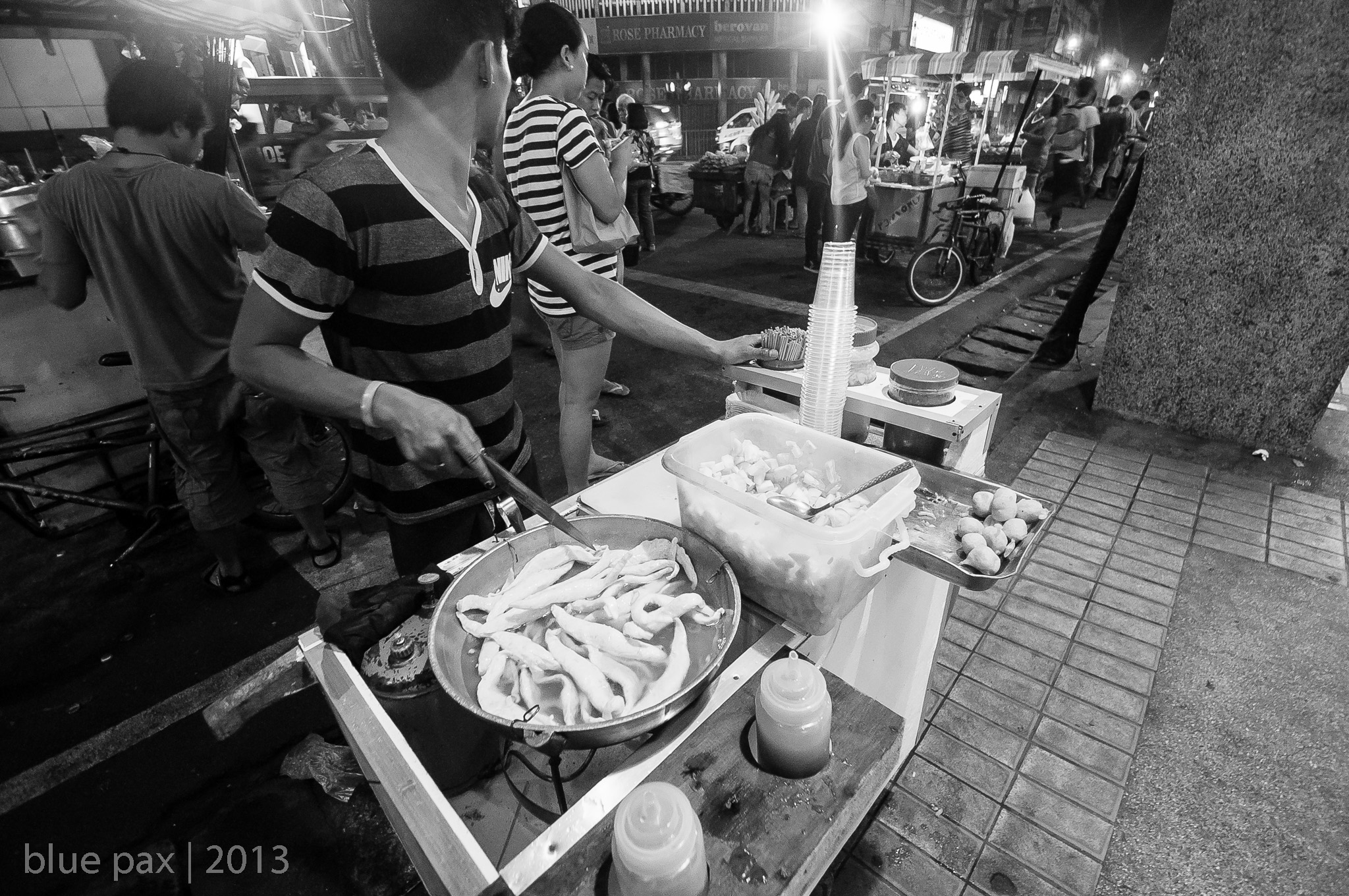 A black-and-white night market scene showing a street food vendor leaning over a stall, preparing food with tongs while bowls of ingredients sit on a makeshift wooden counter.