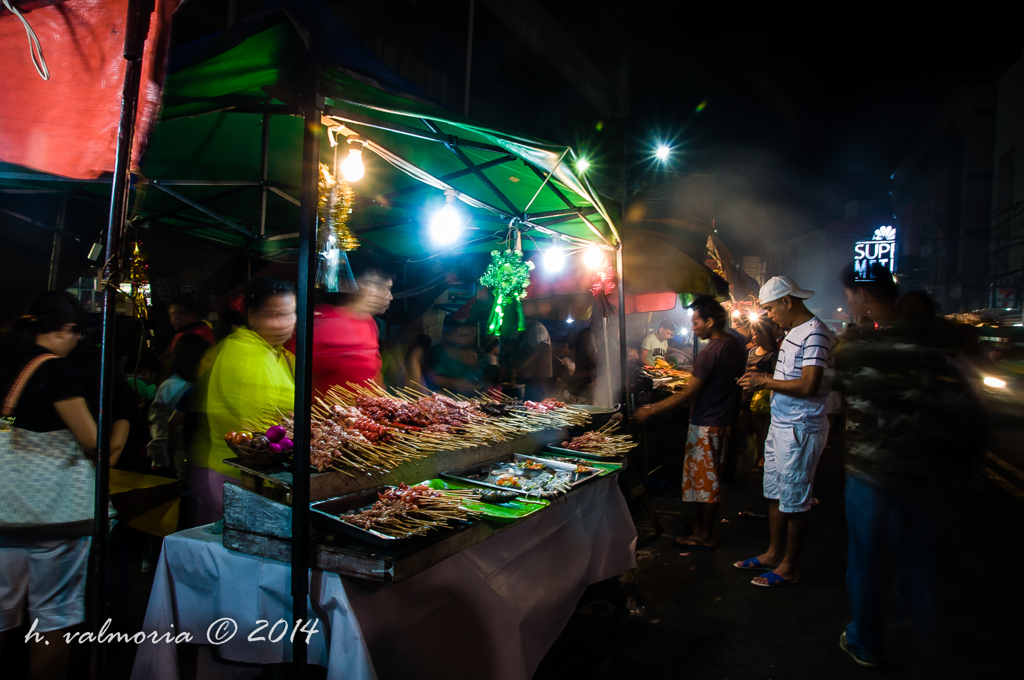 A bustling outdoor night market food stall illuminated by hanging bulbs, with rows of meat skewers displayed on a table while vendors in colorful shirts serve customers gathered in the dark street.