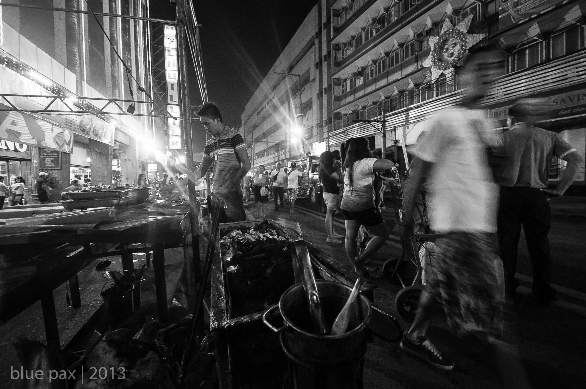 A black-and-white night photograph of a busy urban street market, with a vendor manning a grill in the foreground and crowds of pedestrians lit by dramatic lens flare from streetlights.