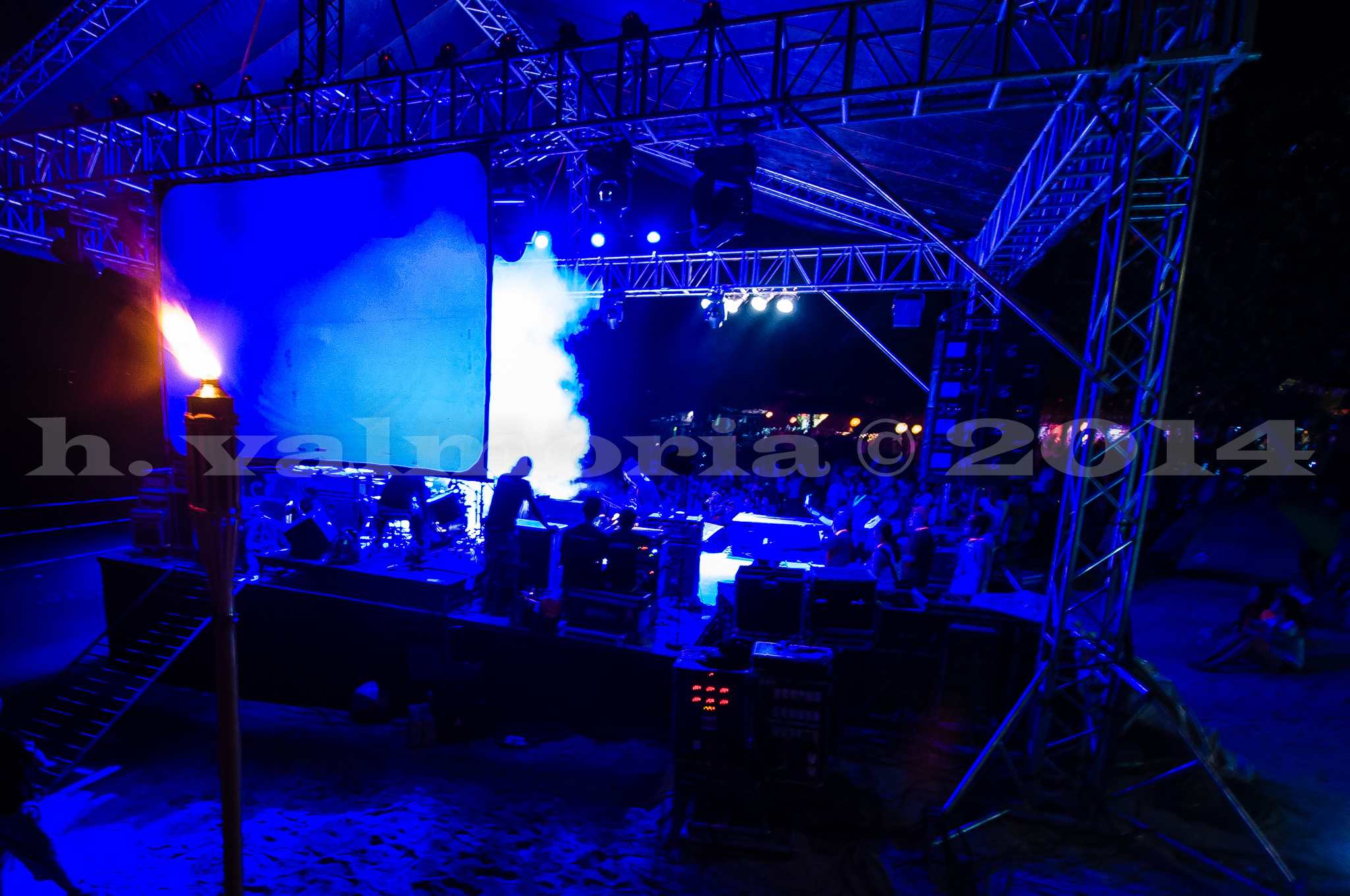 An outdoor concert stage bathed in deep blue lighting with smoke rising from behind a large projection screen, while a fire torch burns on the left side of the frame and rigging trusses frame the scene from above.