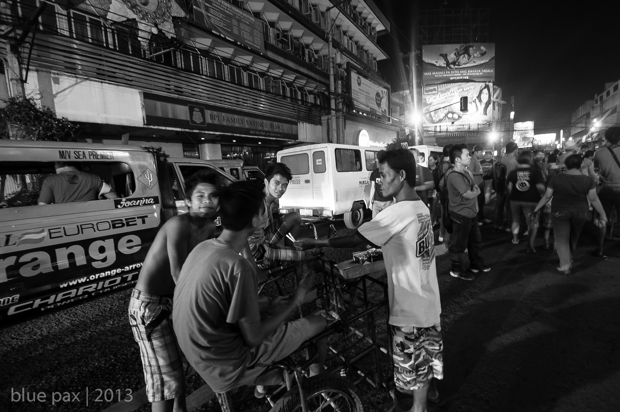 Several young Filipino men gathered around a pedicab on a busy urban street at night in black and white, with a jeepney, buildings, and billboards visible in the background.