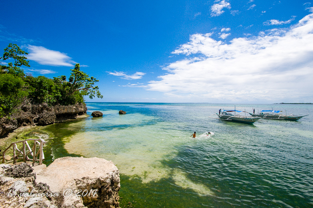 A vibrant Philippine coastal cove with clear turquoise shallows, rocky limestone outcroppings, and lush green trees, while a traditional outrigger bangka boat floats offshore under a bright blue sky. A lone swimmer wades in the calm water.