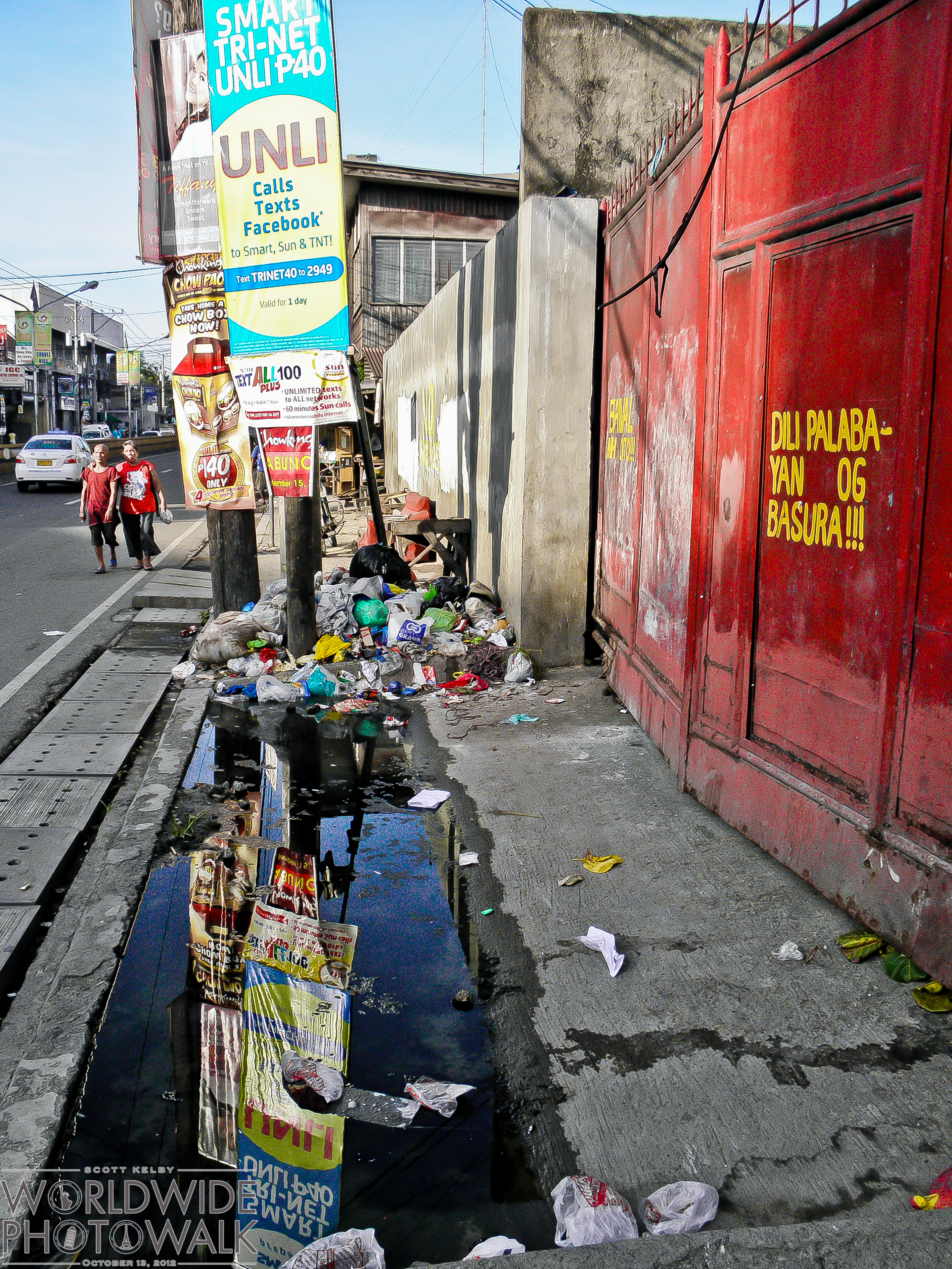 A cluttered Philippine city sidewalk with an open drainage canal filled with garbage and stagnant water, while a red wall bears a Cebuano warning against littering.