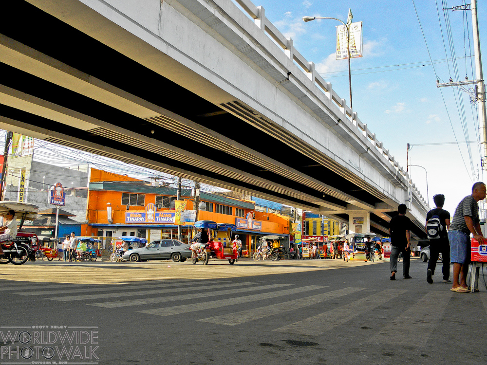 A busy Philippine street intersection beneath a concrete elevated highway overpass, with colorful shopfronts, tricycles, and pedestrians crossing under a blue sky.
