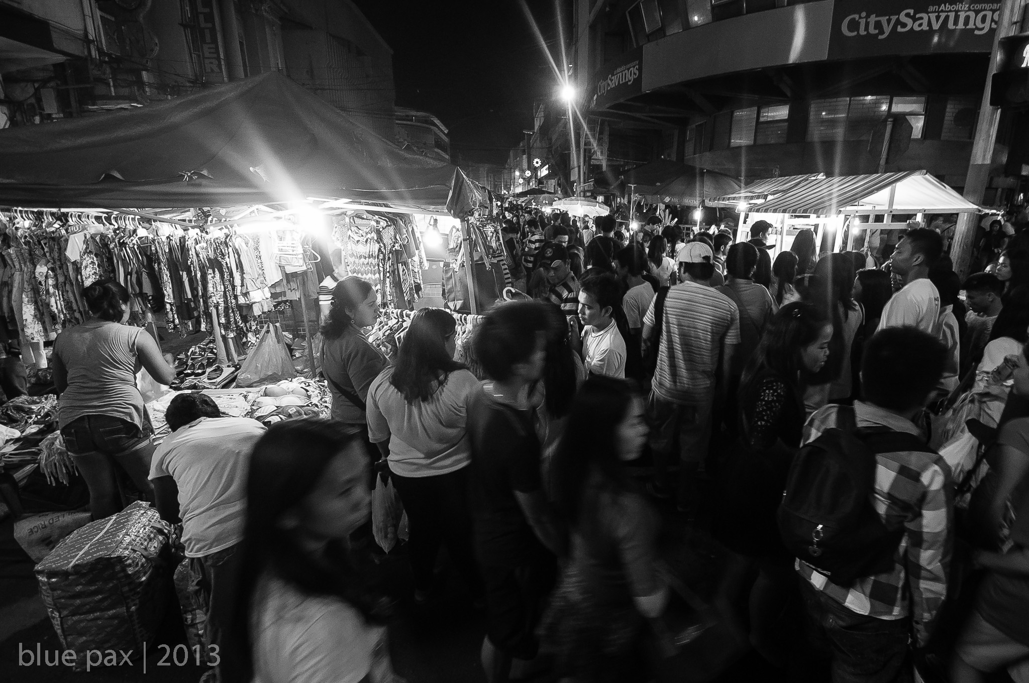 A dense crowd of shoppers flowing through a bustling night market in the Philippines in black and white, with clothing stalls illuminated by starburst streetlights lining the busy street.