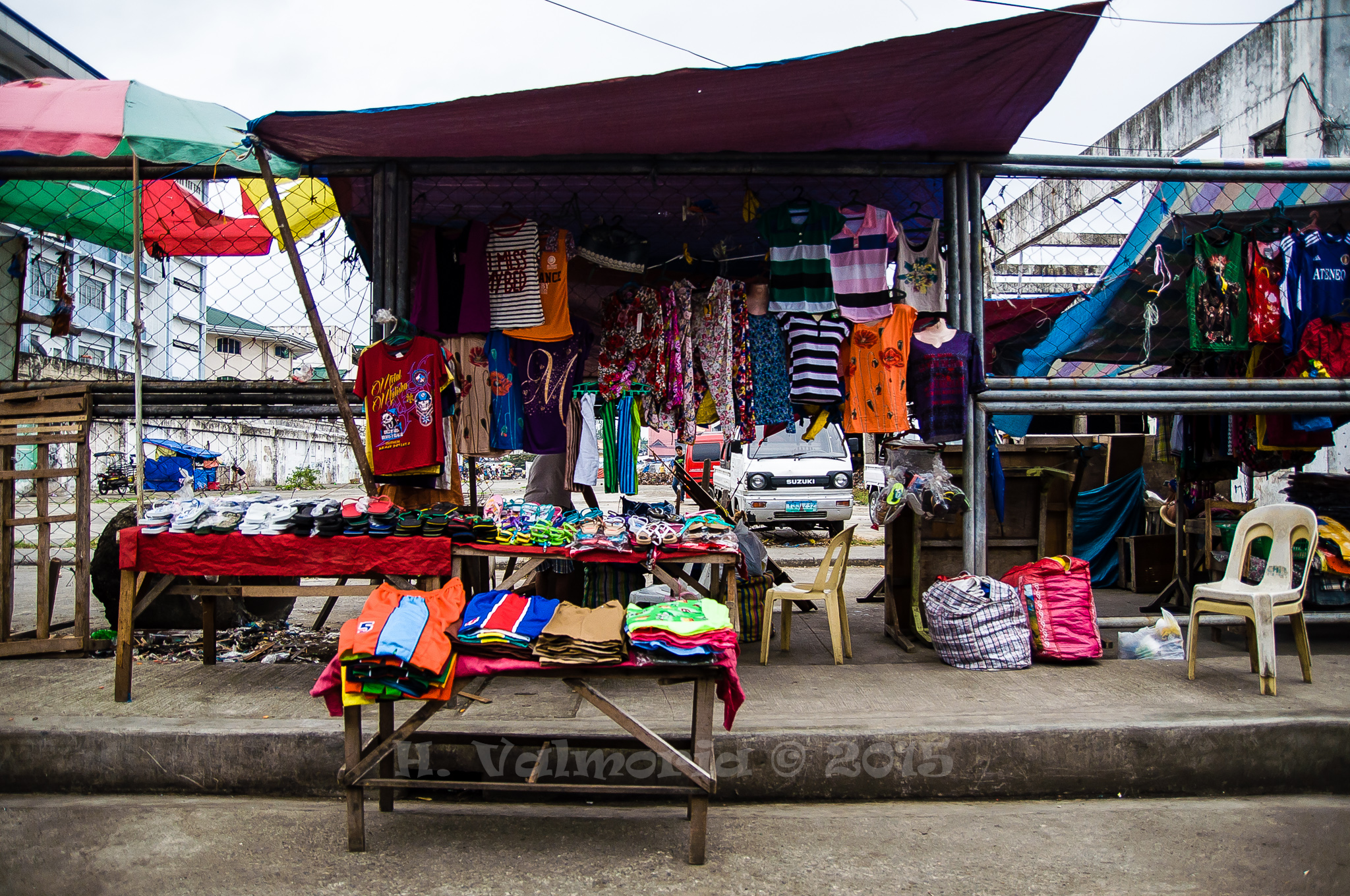 A colorful roadside market stall displays hanging clothing, scarves, and stacked goods on wooden tables beneath tarpaulin awnings, with a chain-link fence and concrete building in the background.