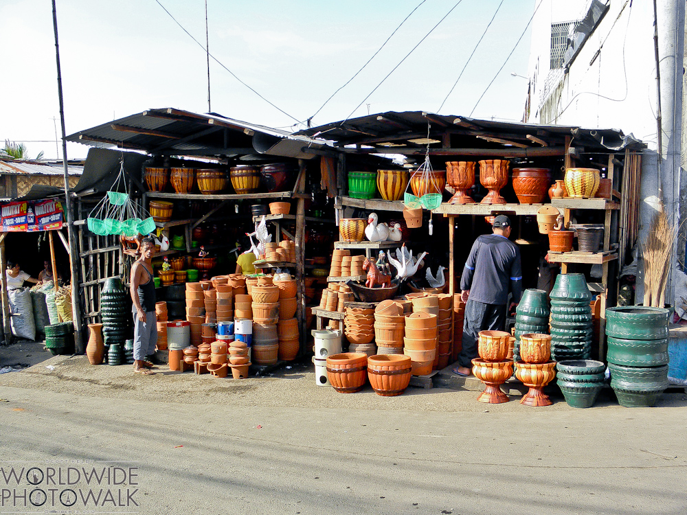 A roadside market stall displays rows of terracotta and green glazed pots, urns, and ceramic planters under a corrugated metal canopy, with two vendors standing among the wares.
