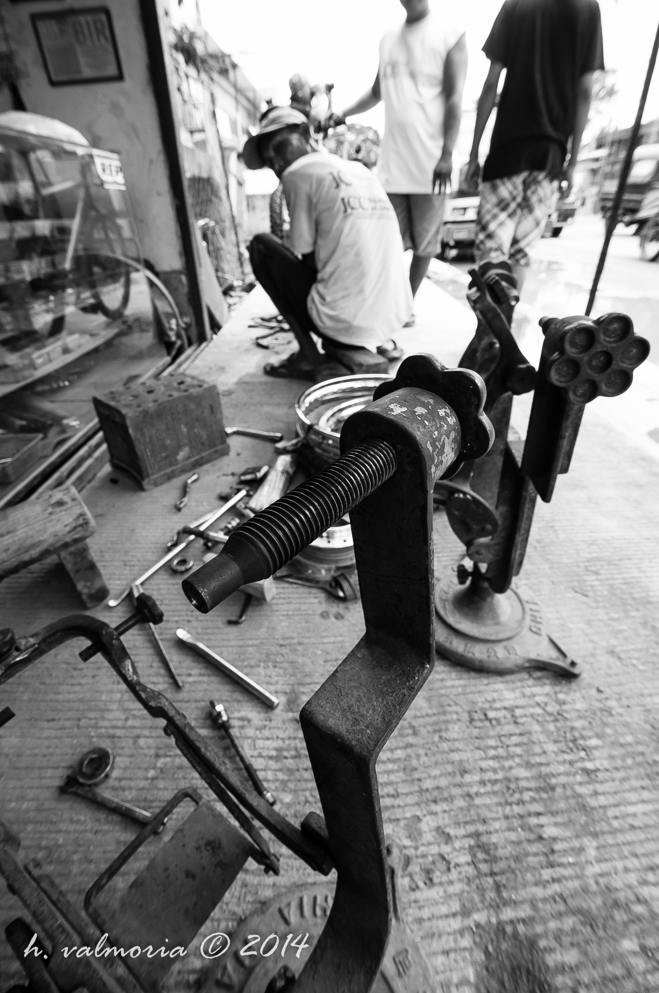 A street mechanic crouching on a sidewalk surrounded by metal tools and machinery parts in black and white, with pedestrians and a busy street visible in the background.