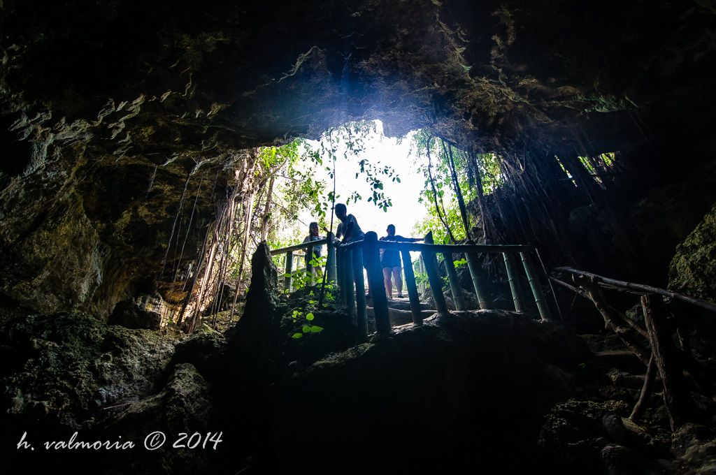 Silhouetted figures stand on a wooden walkway at the entrance of a dramatic cave, framed by jagged rock formations and lush tropical vegetation with bright light flooding in from outside.