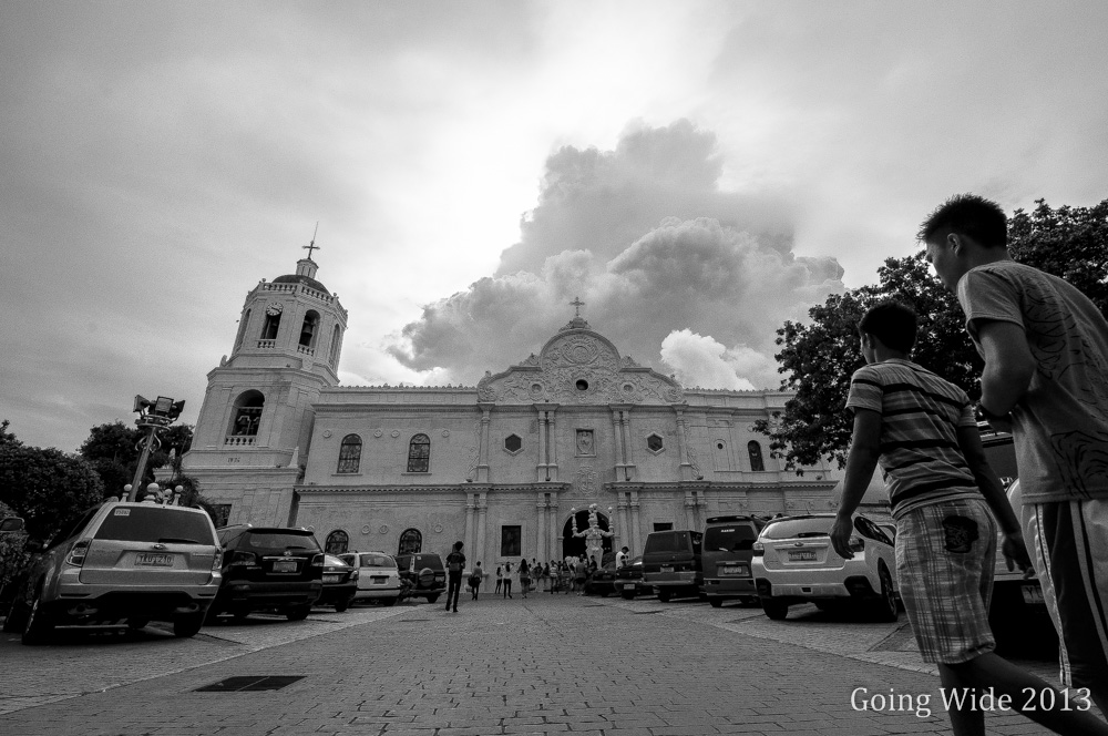 Two young men walk past a grand Spanish colonial church in black and white, its ornate baroque facade rising beneath dramatic storm clouds, with parked cars and churchgoers filling the cobblestone plaza.