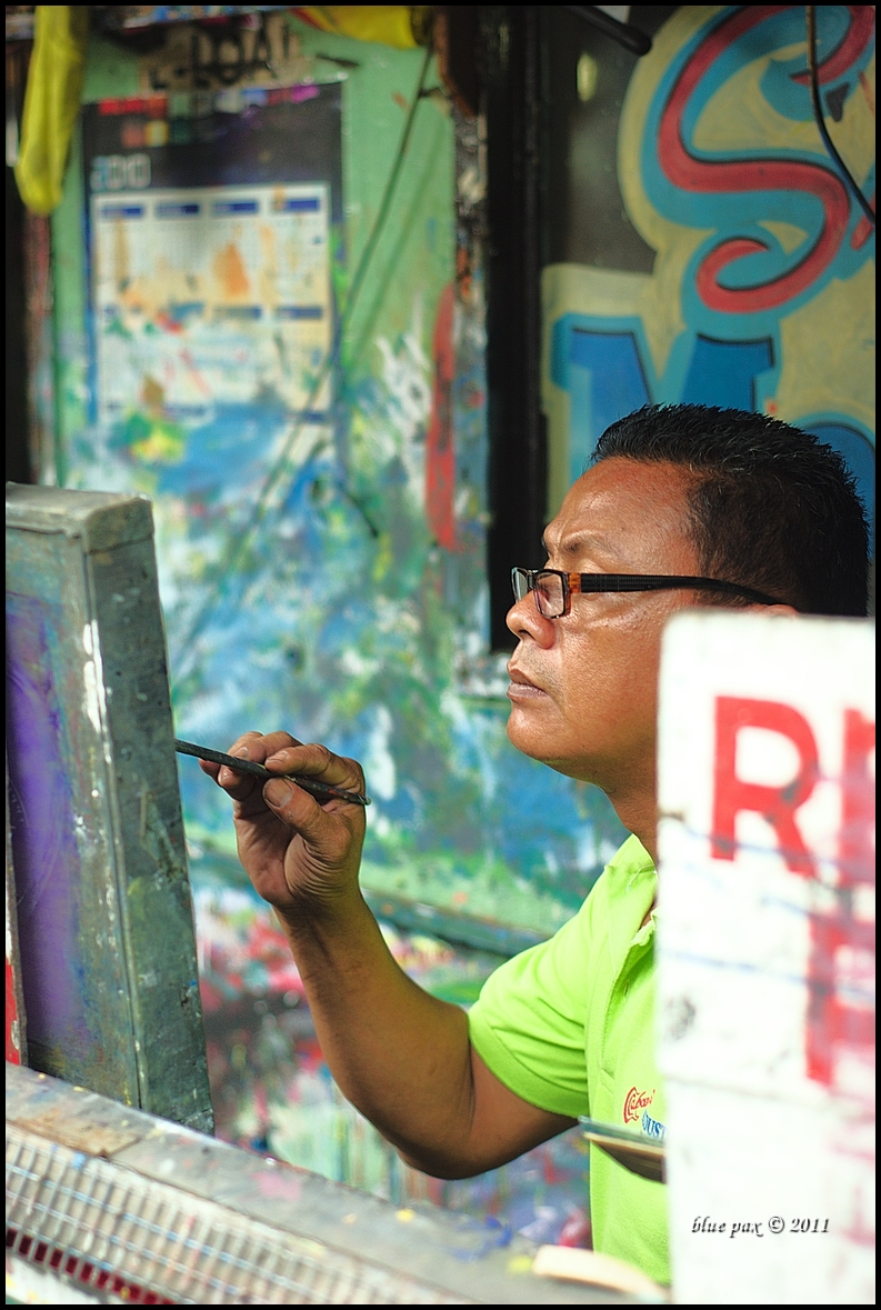 A man wearing glasses and a lime green shirt concentrates as he applies fine brushstrokes to a canvas in a paint-splattered street workshop, surrounded by colorful signage and graffiti.