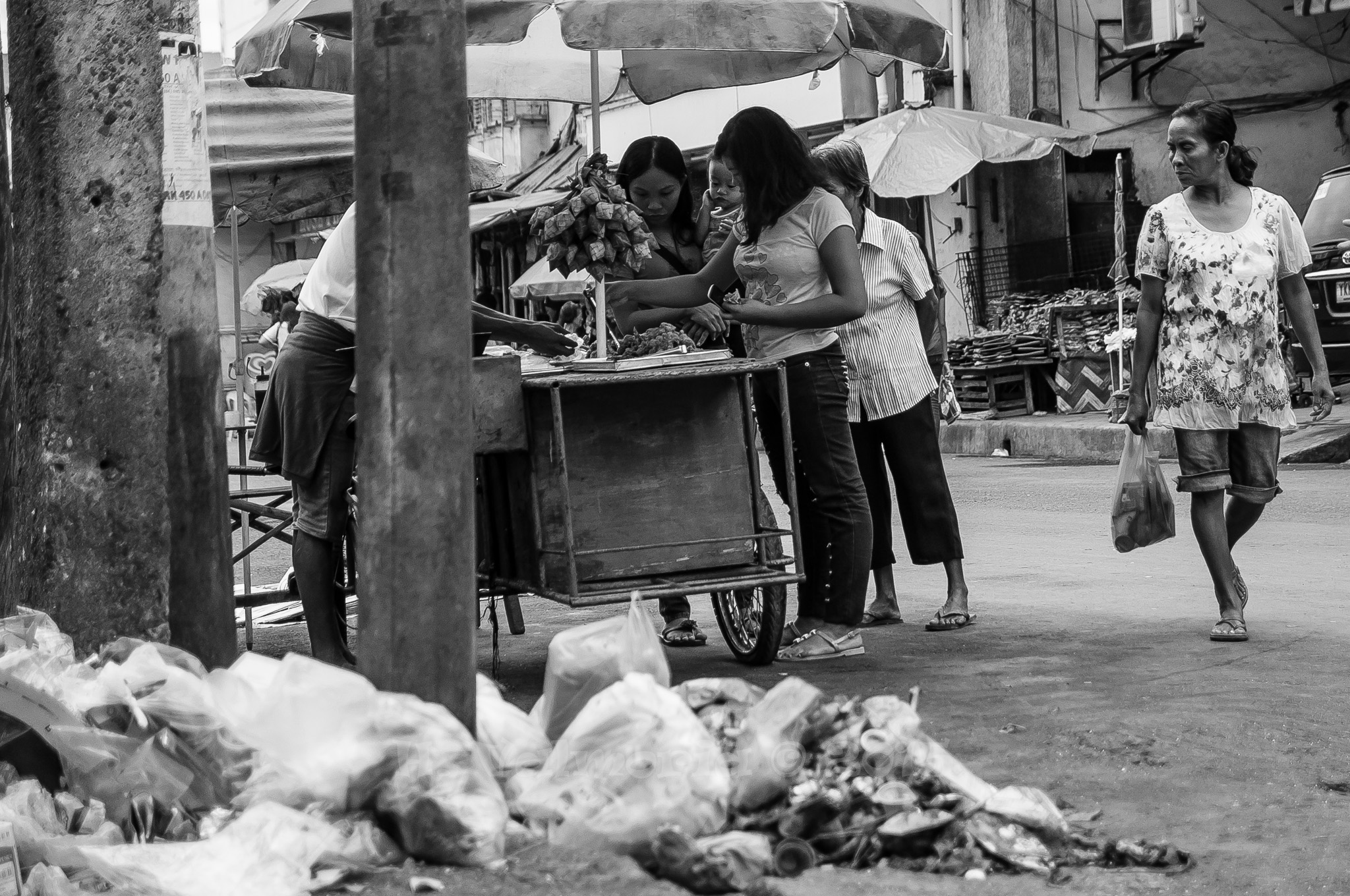 Two women browse a street vendor cart in a busy Southeast Asian market in black and white, while a third woman walks past, with garbage bags visible in the foreground.