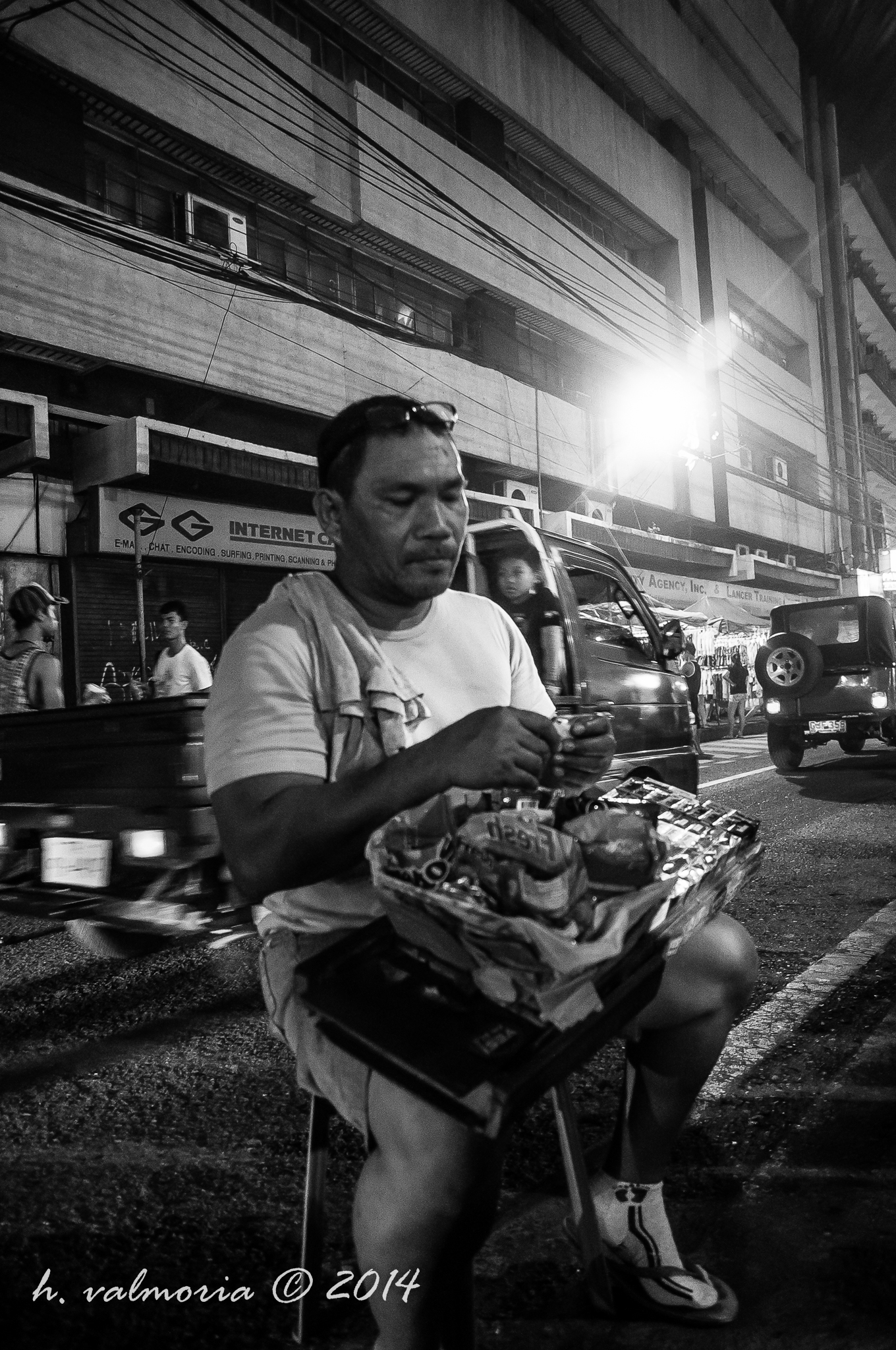 A man seated on a small stool along a busy urban street at night, holding a tray of small goods for sale, with traffic and shopfronts blurred in the background, in black and white.