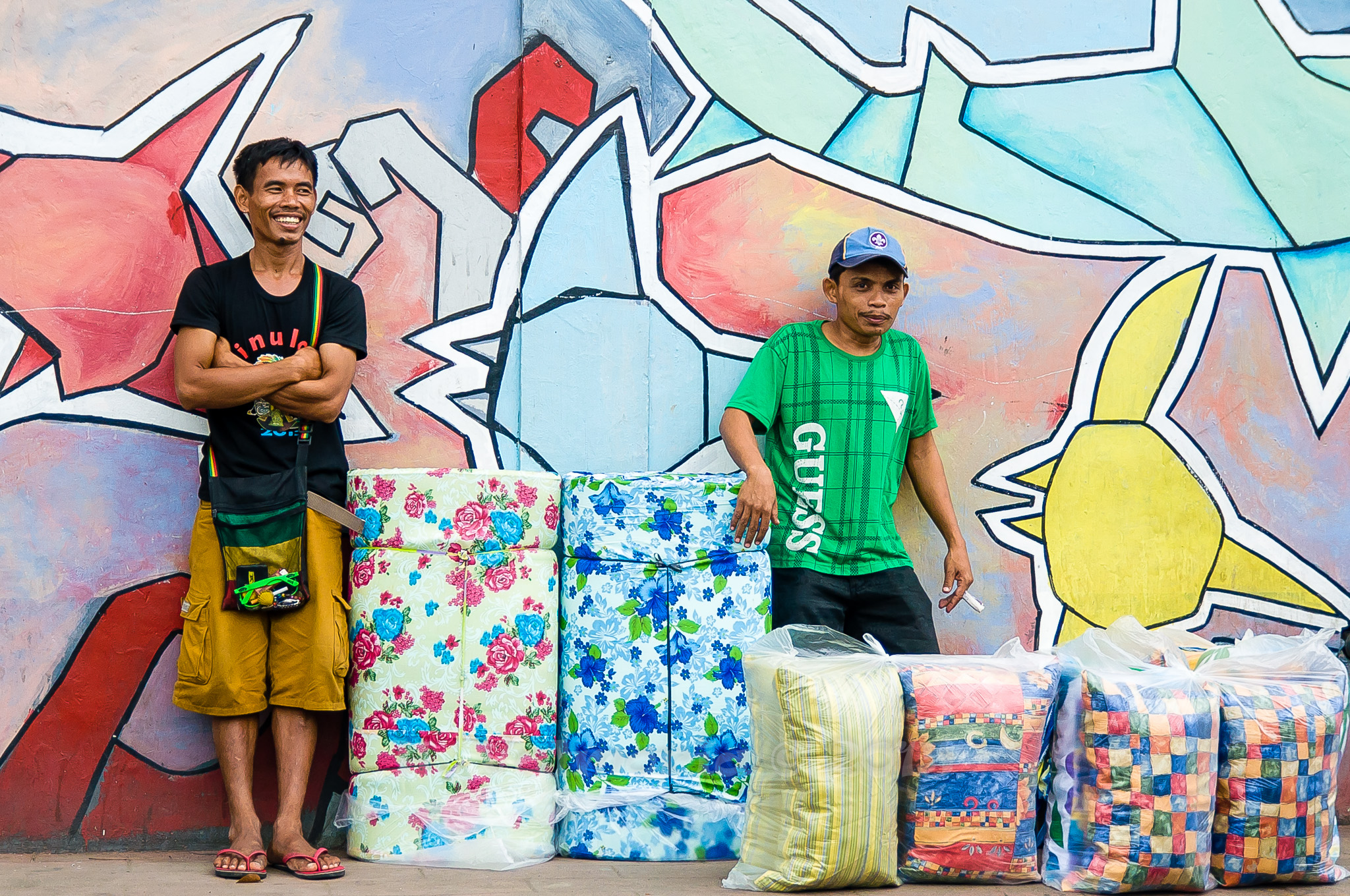 Two male street vendors stand in front of a bold, colorful graffiti mural, their patterned bedding goods arranged on the ground between them, one man smiling with arms crossed.