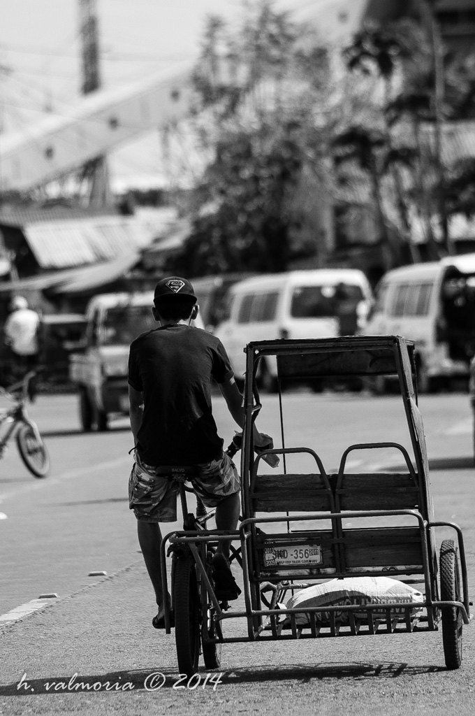 A young Filipino man pedals a traysikad away from the camera through a busy urban street in high-contrast black and white, with traffic and market stalls blurred in the background.