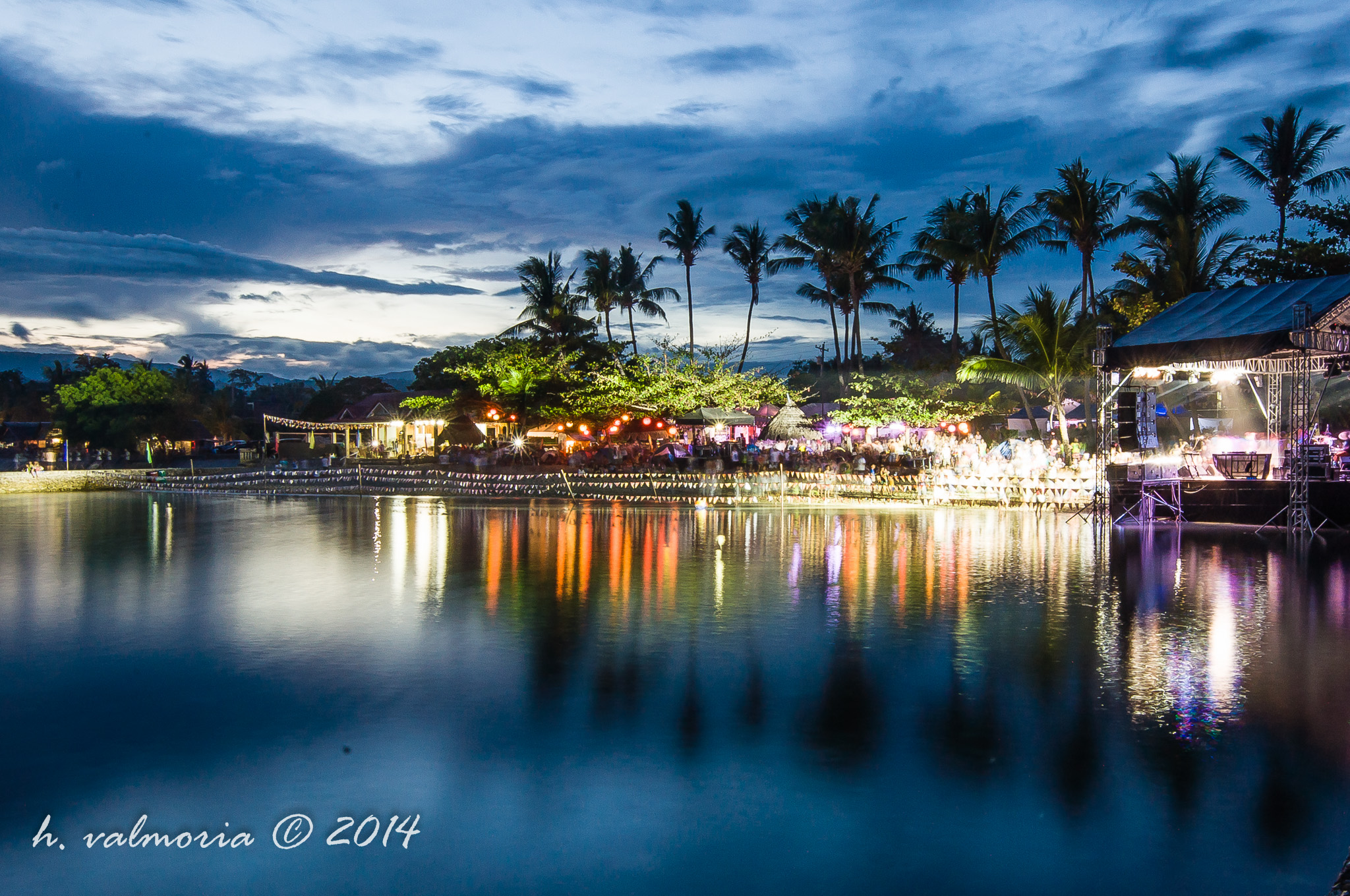 A twilight scene of a festive outdoor venue along a calm reflective waterway in the Philippines, with warm event lights glowing among palm trees silhouetted against a deep blue cloudy sky.